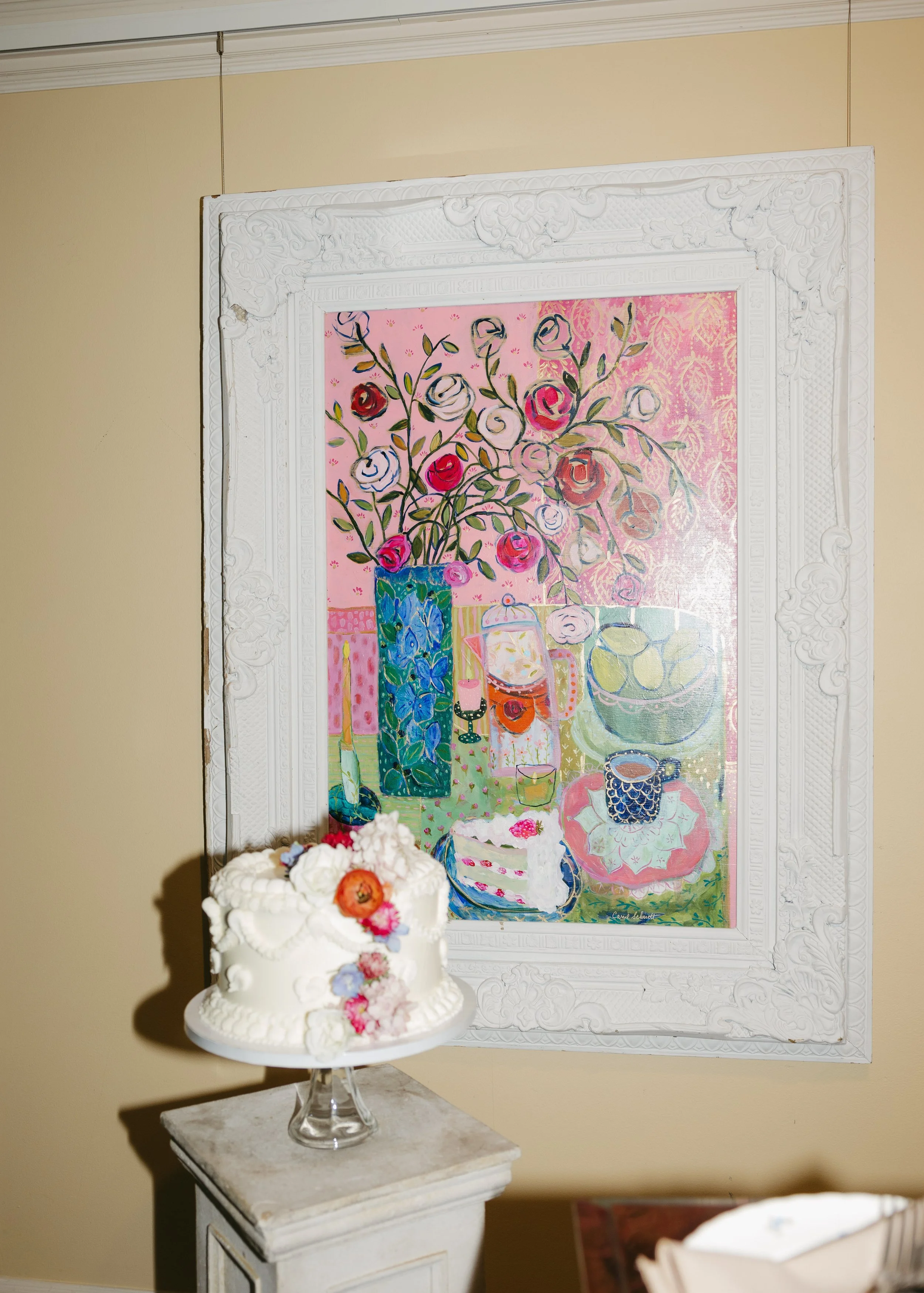 A white decorated cake with flowers on top, placed on a small white table, positioned in front of a colorful painting of a still life with flowers and tableware. The painting has a pink background with various objects and flowers, and is framed with 