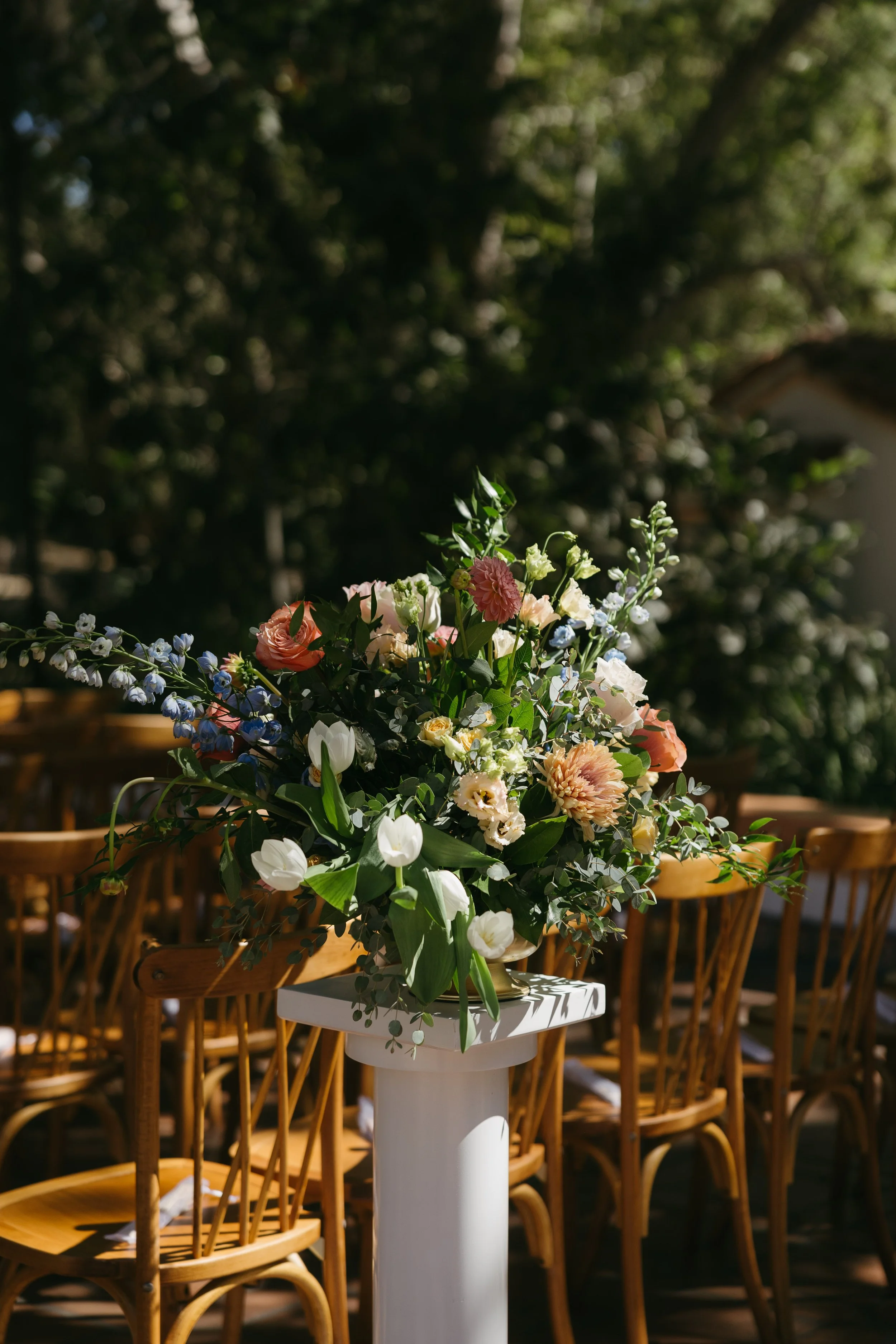A large floral arrangement with pink, white, and blue flowers on a white pedestal in an outdoor setting with wooden chairs nearby.