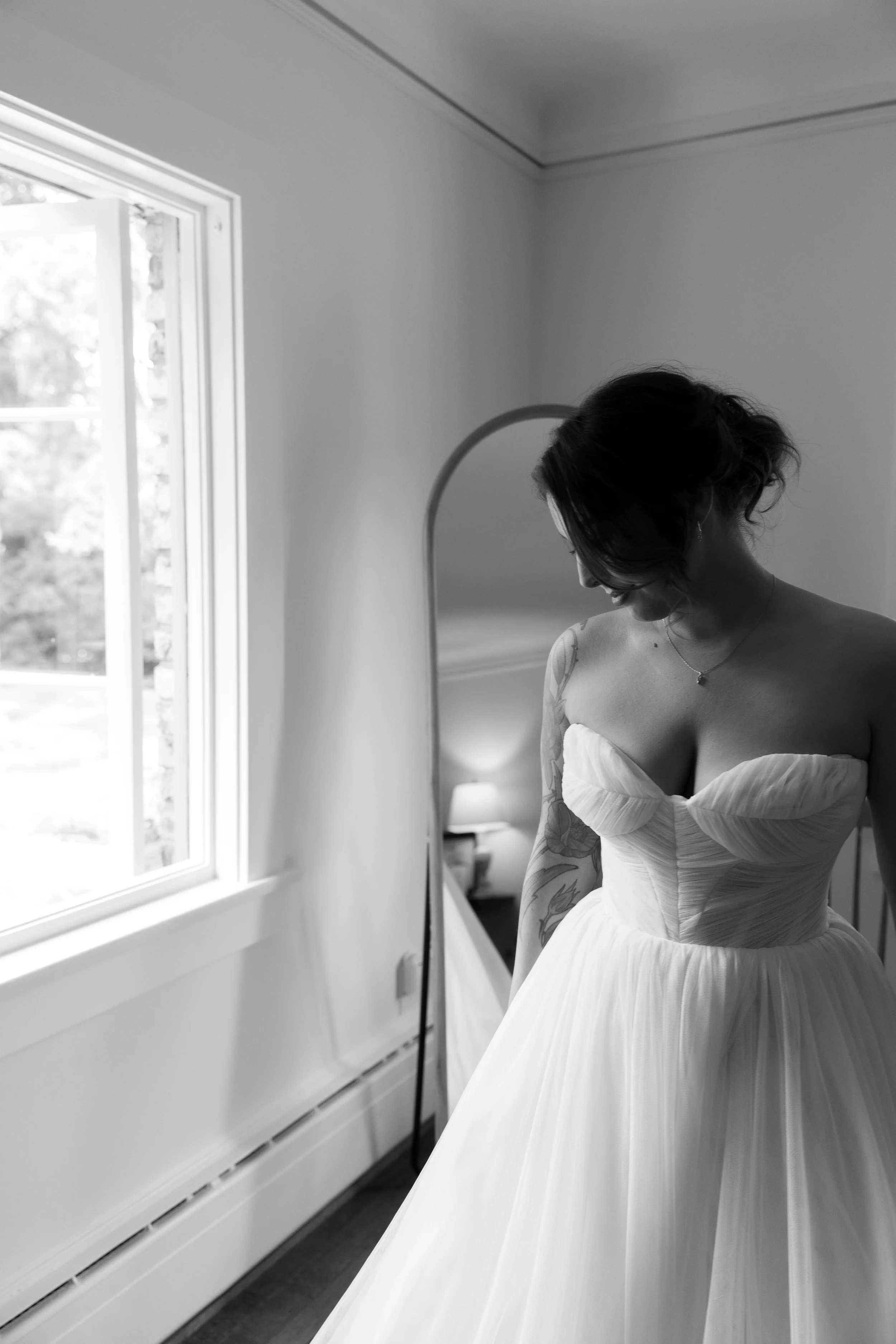 A woman in a strapless wedding dress looking down in a room with a window, mirror, and lamp.