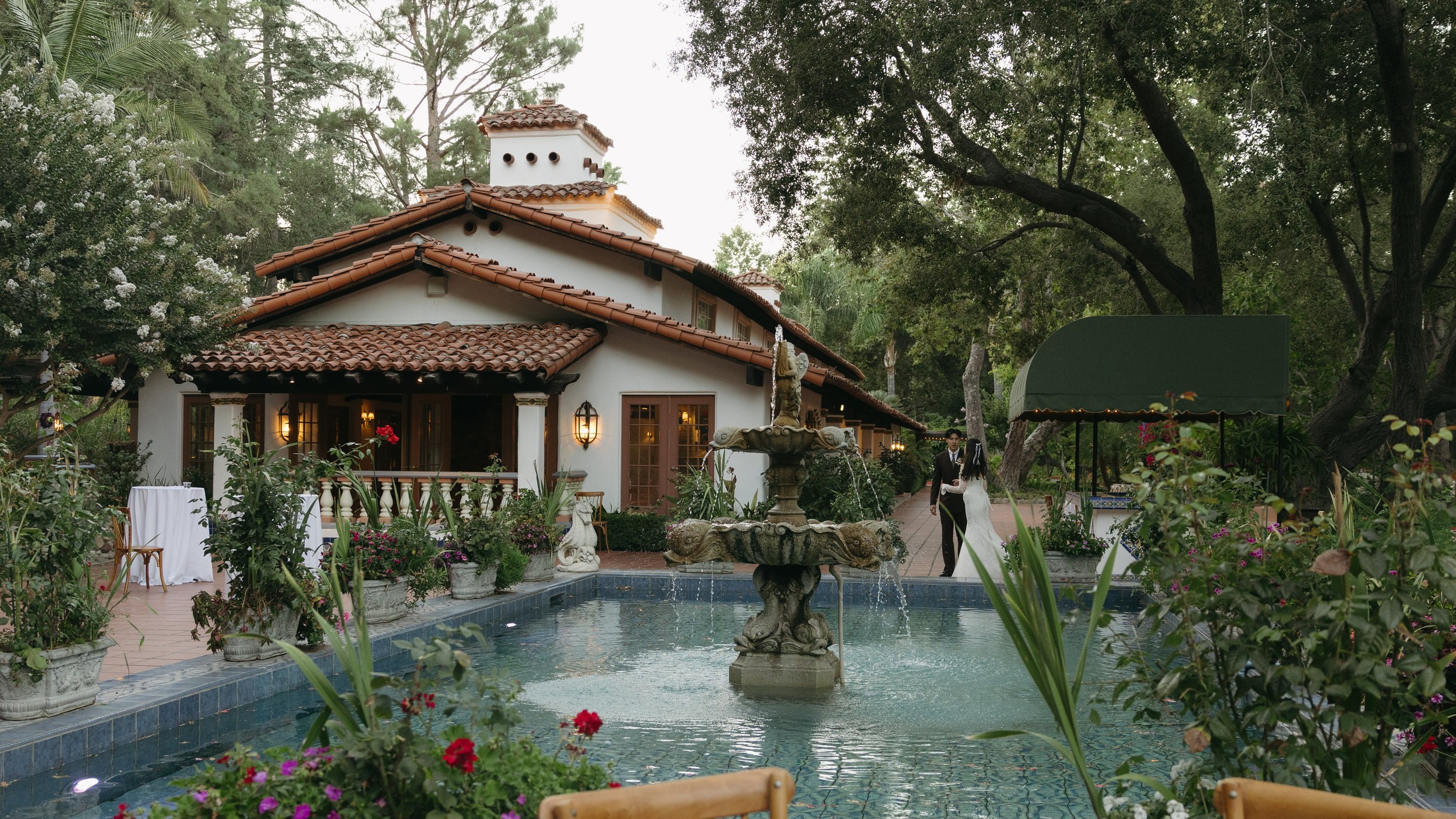 A wedding couple walking beside a fountain in a garden with a scenic house in the background at a Rancho Las Lomas Wedding in Los Angeles