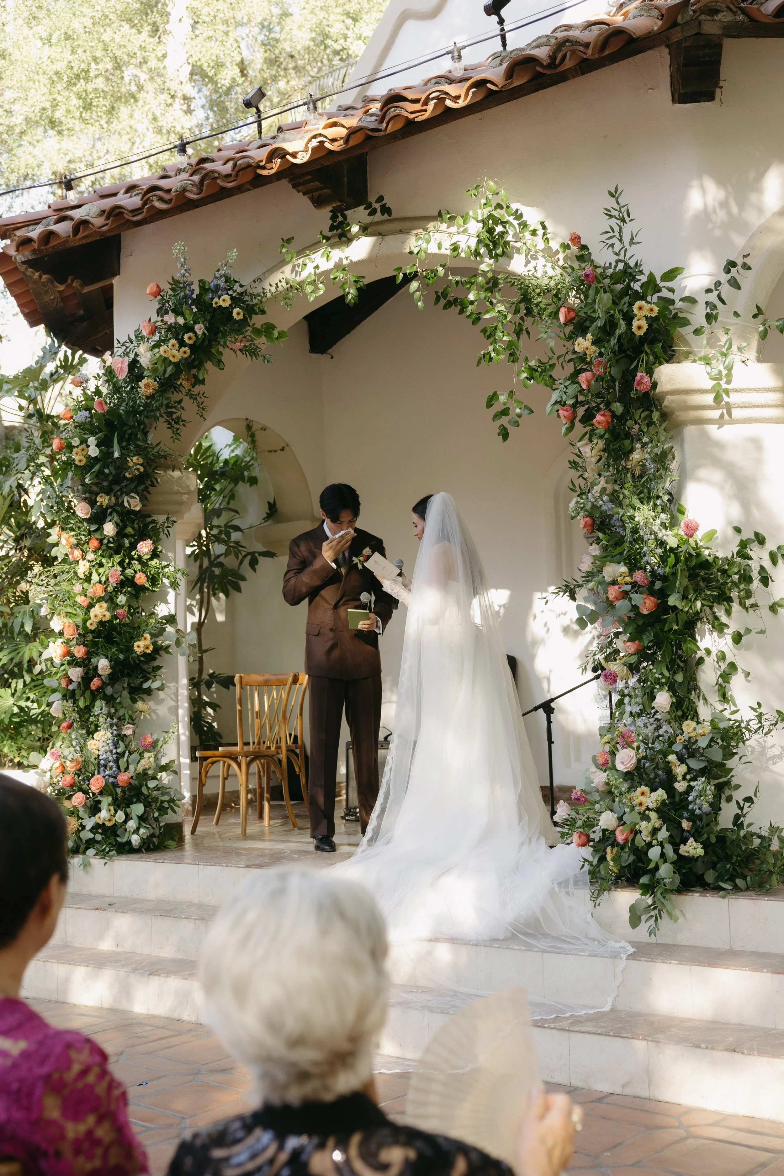 A bride and groom exchanging vows during a wedding ceremony outdoors under a floral arch with guests watching.