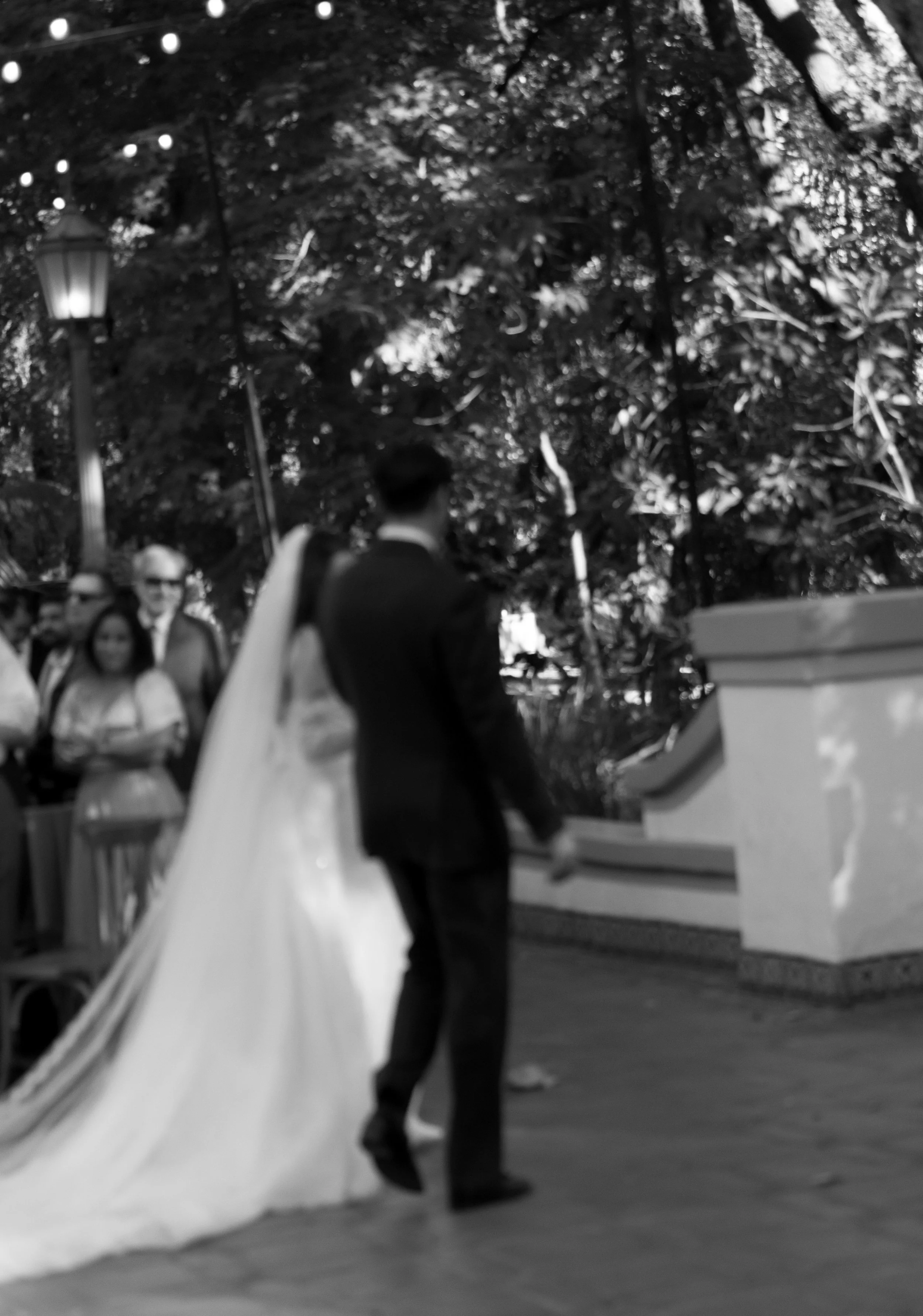 A black-and-white photo of a wedding ceremony outdoors with a bride and groom, surrounded by guests, trees, and a lamppost at a Rancho Las Lomas Wedding in Los Angeles
