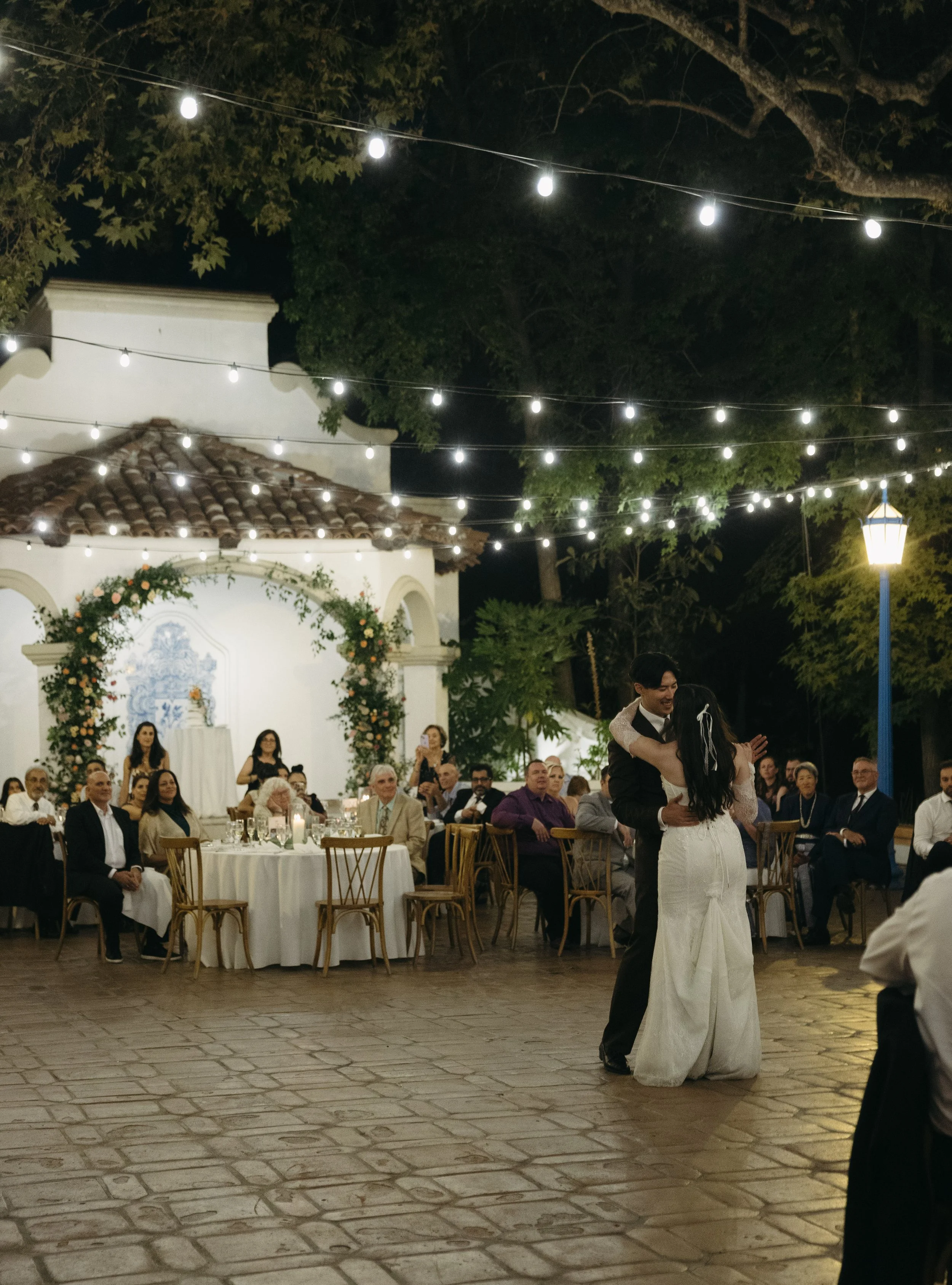 A couple dancing at their wedding reception outdoors at night, with guests seated at tables in the background and string lights overhead.