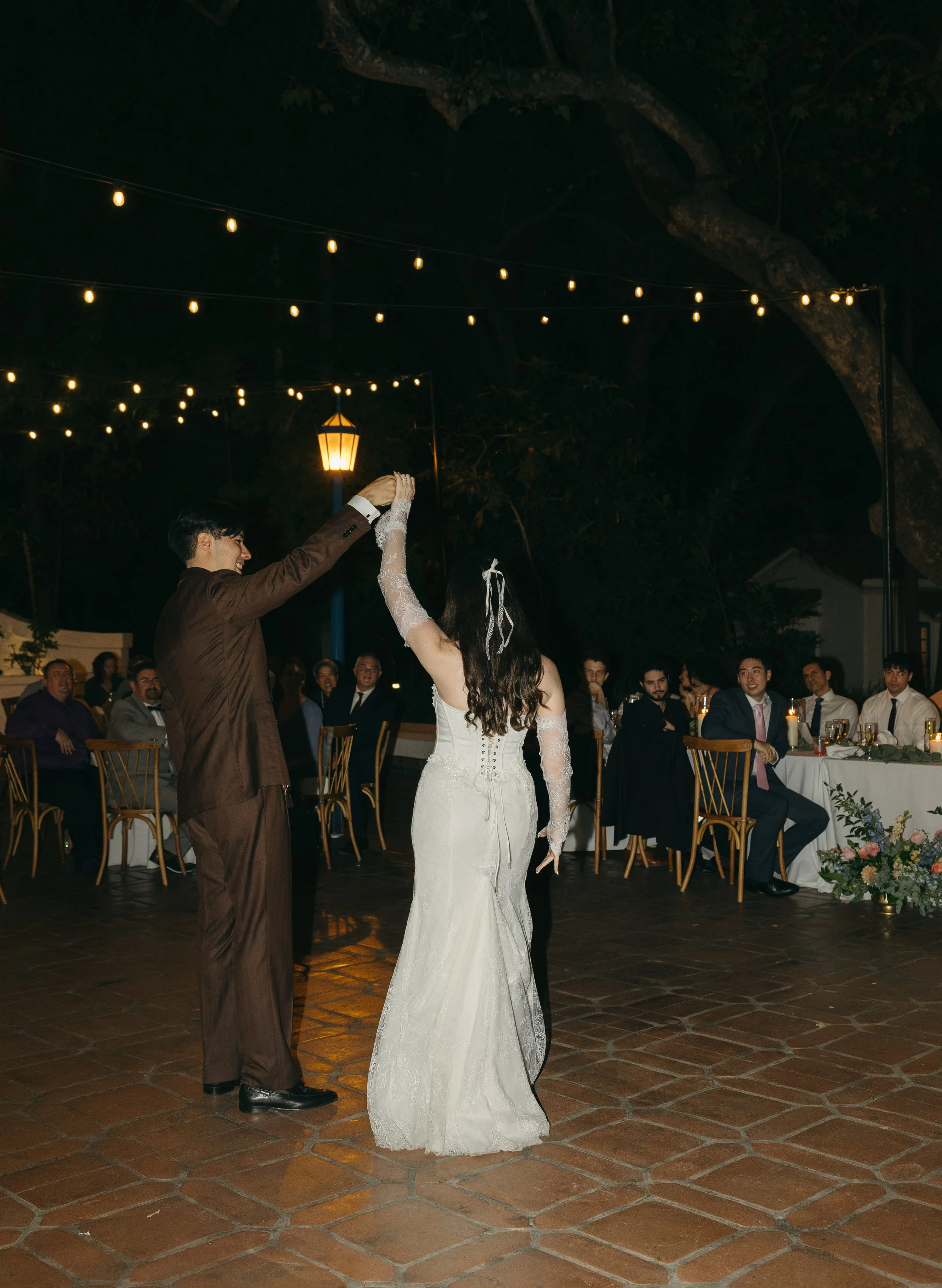 A newlywed couple dances at their outdoor wedding reception at night, with guests seated at tables watching under string lights and a tree overhead.