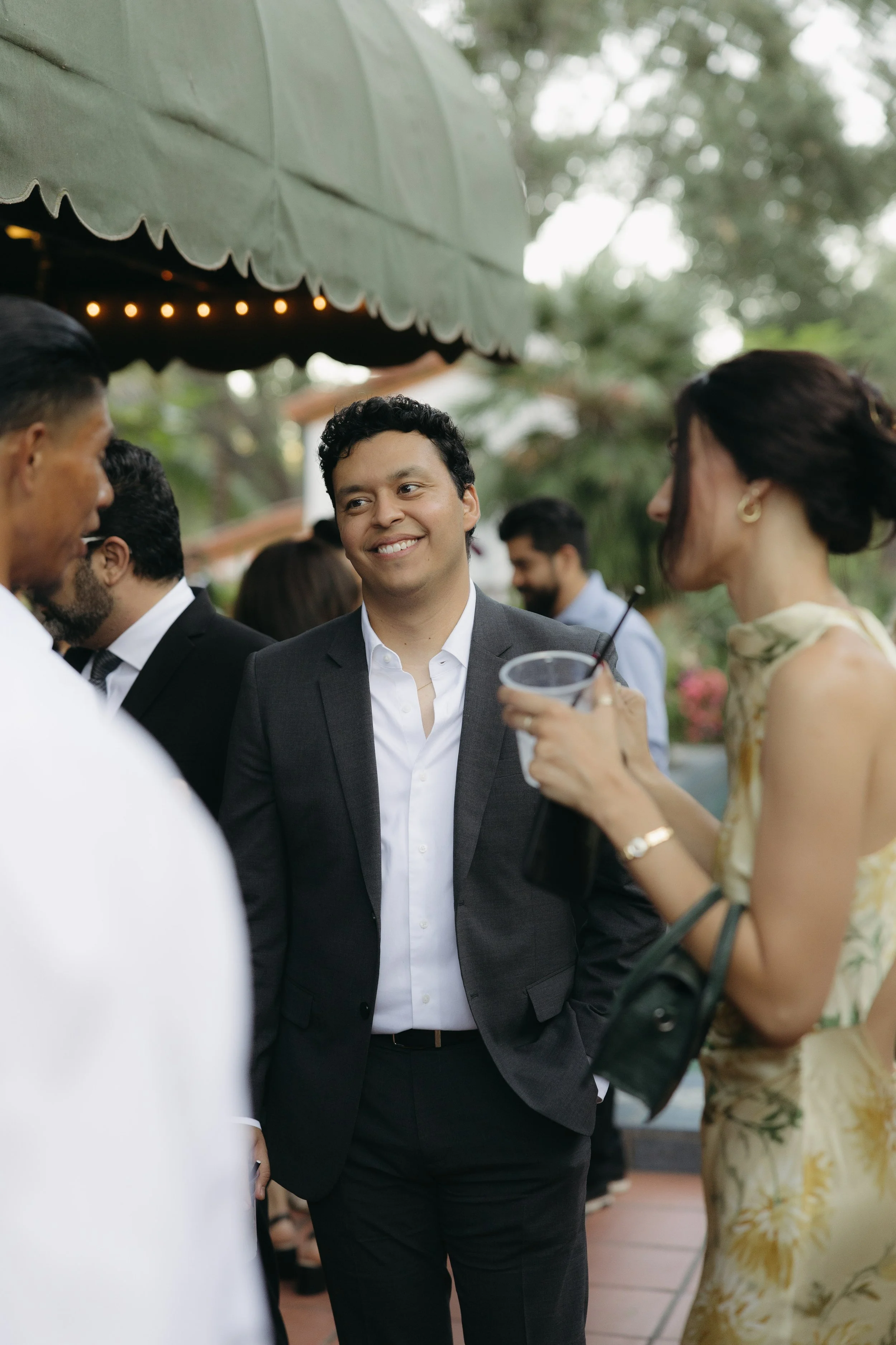 People socializing outdoors at a garden party, with a man in a gray suit smiling and woman holding a drink.