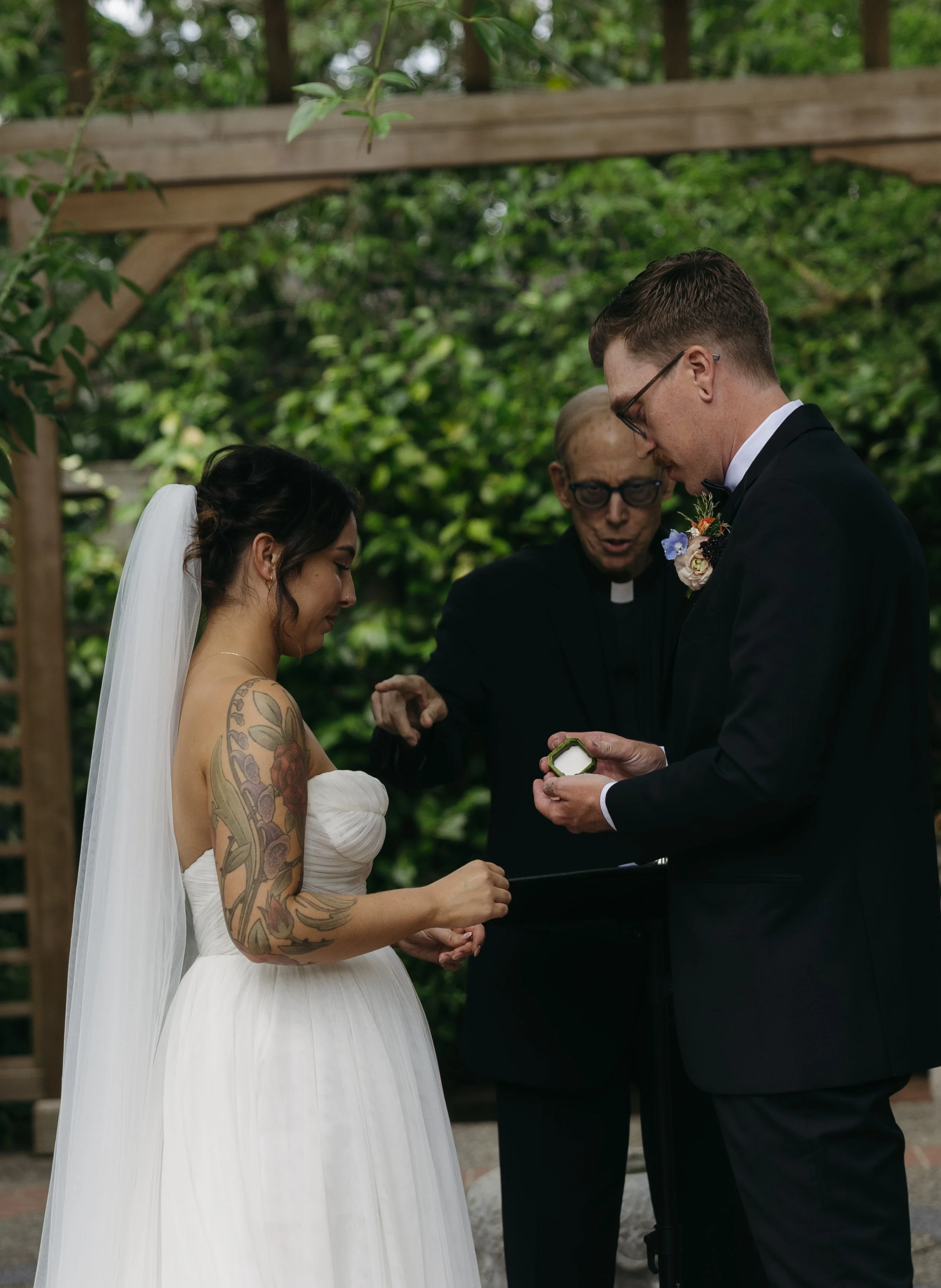 A bride and groom exchanging rings during a wedding ceremony outdoors, officiated by a minister. The bride has tattoos on her arm and is wearing a white strapless wedding gown and veil. The groom is dressed in a black tuxedo with glasses. They are un