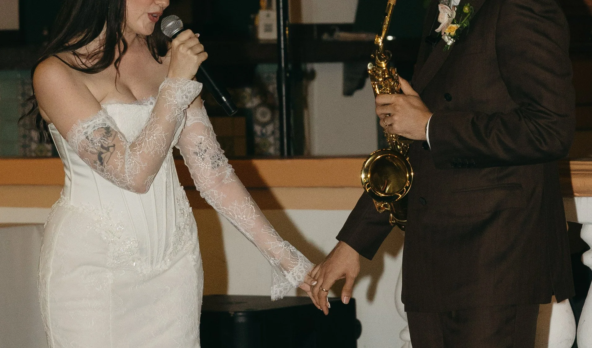 A bride and groom holding hands during their wedding ceremony. The bride is wearing a white lace dress and speaking into a microphone, while the groom, dressed in a dark suit, is holding a gold saxophone.