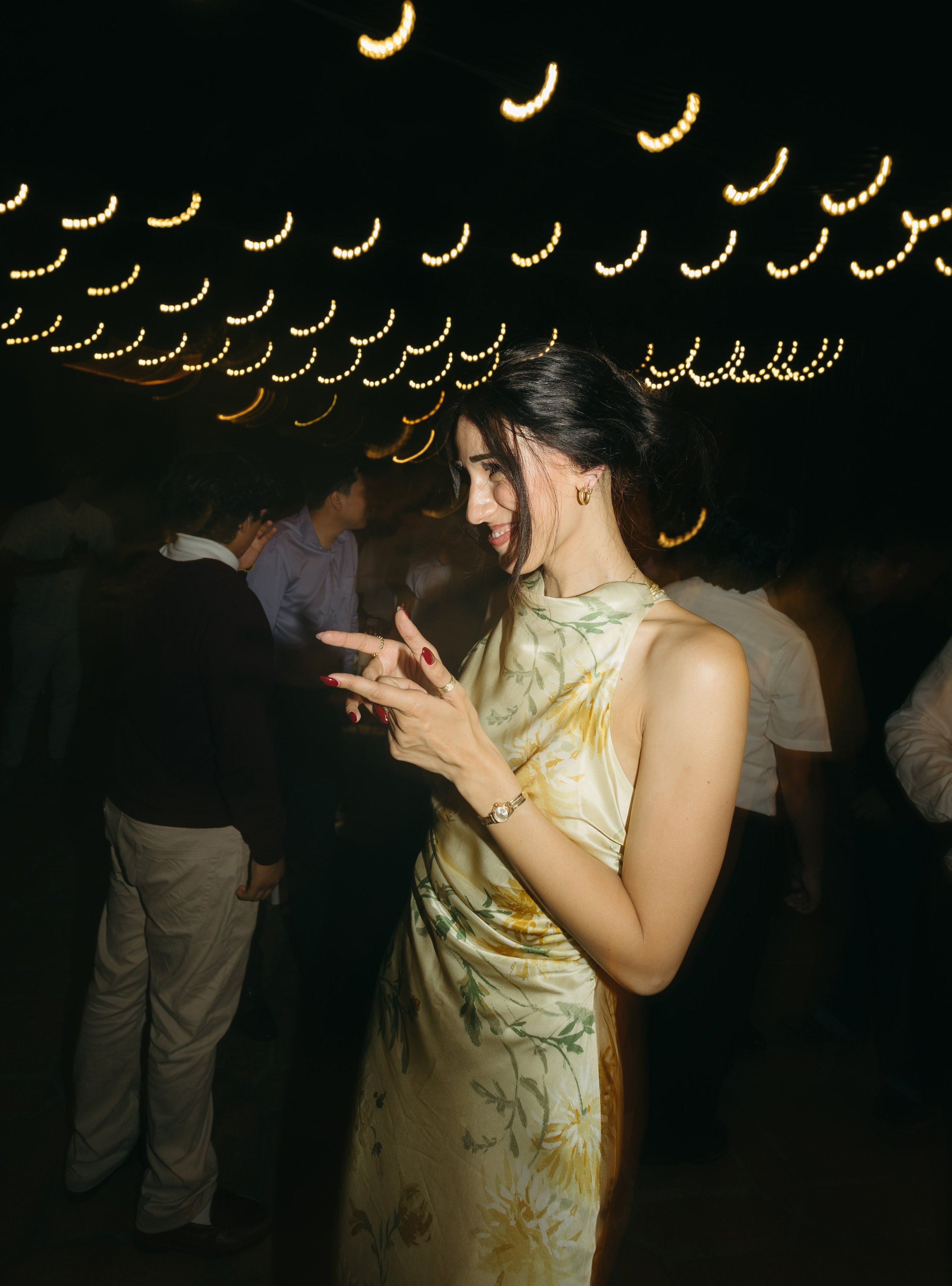 A woman in a floral satin dress smiling and looking at her phone at a party, with other guests in the background under string lights.