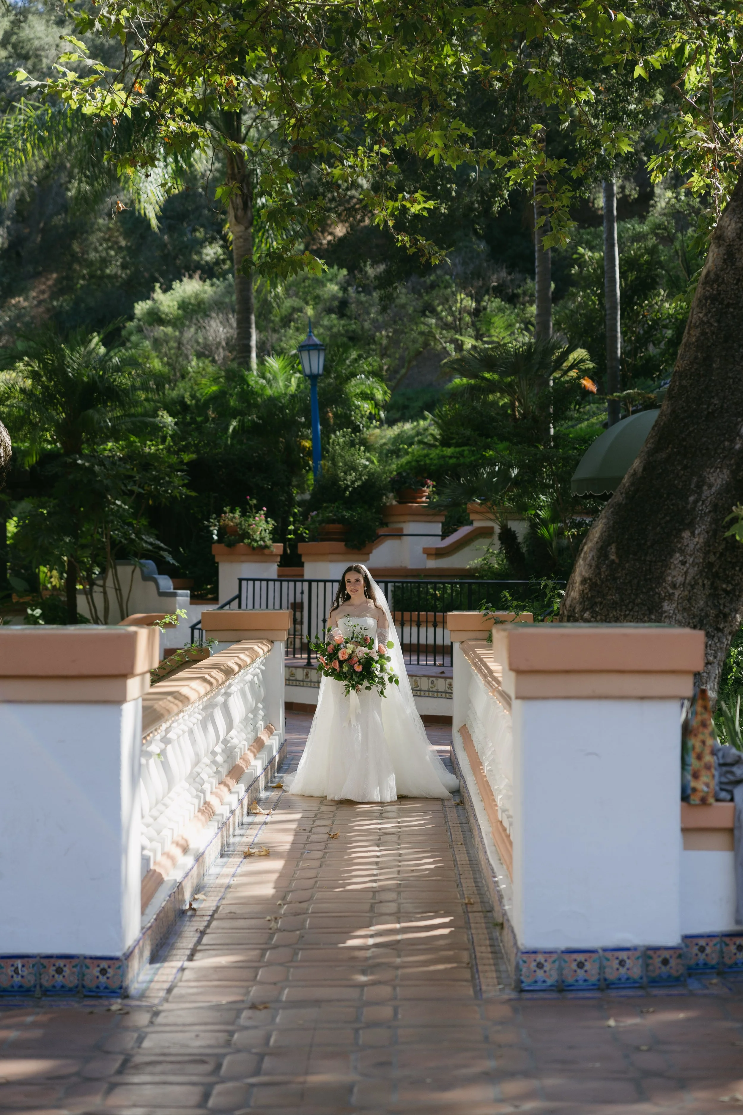 Bride in wedding dress holding a large bouquet of flowers standing on a garden bridge surrounded by lush greenery and trees at a Rancho Las Lomas Wedding in Los Angeles