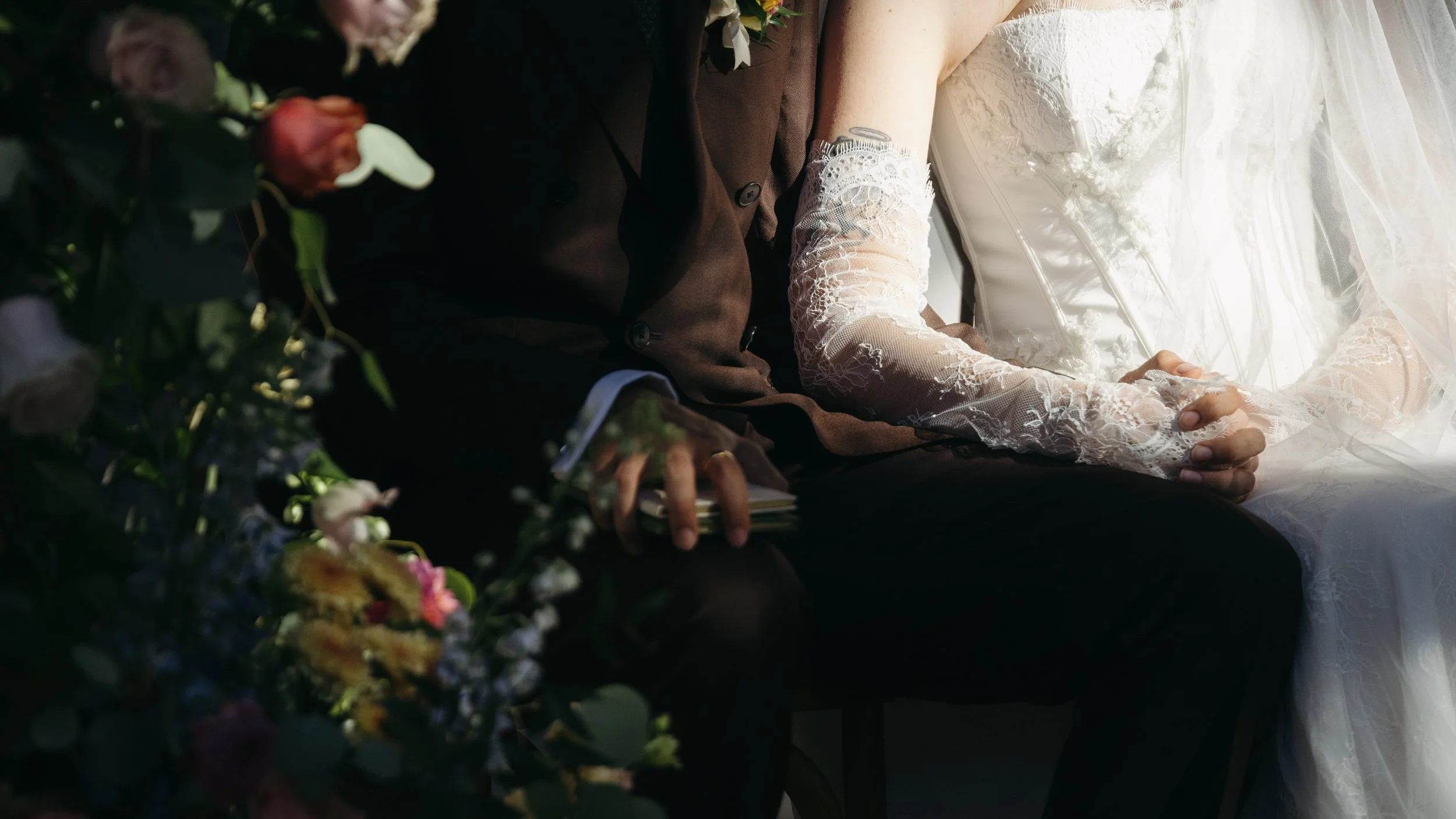 Close-up of a bride and groom sitting together, with the bride wearing a white lace dress and veil, and the groom dressed in a dark suit, surrounded by flowers.