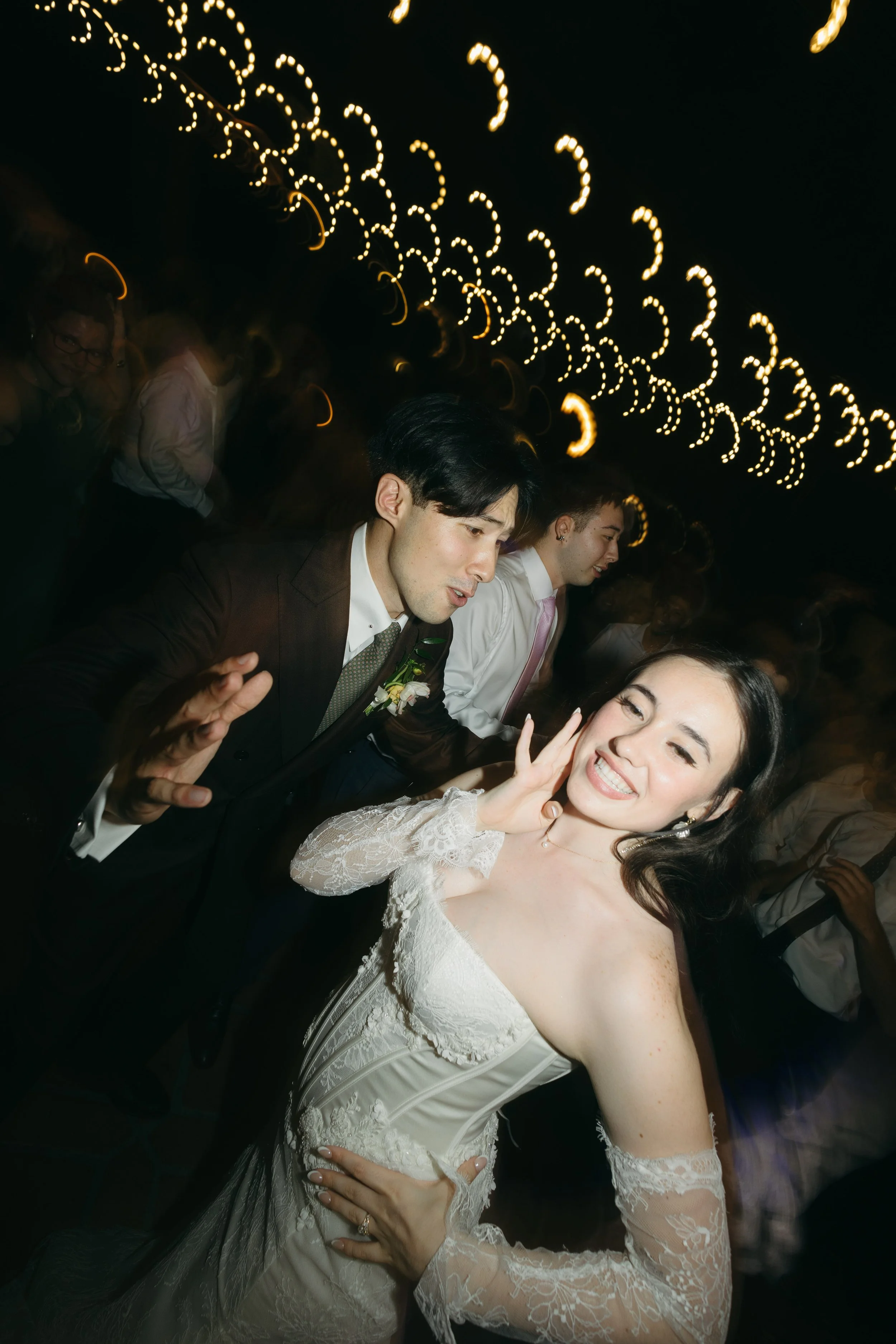A smiling bride in a wedding dress making a peace sign while dancing at her wedding reception with guests in the background and decorative string lights overhead.
