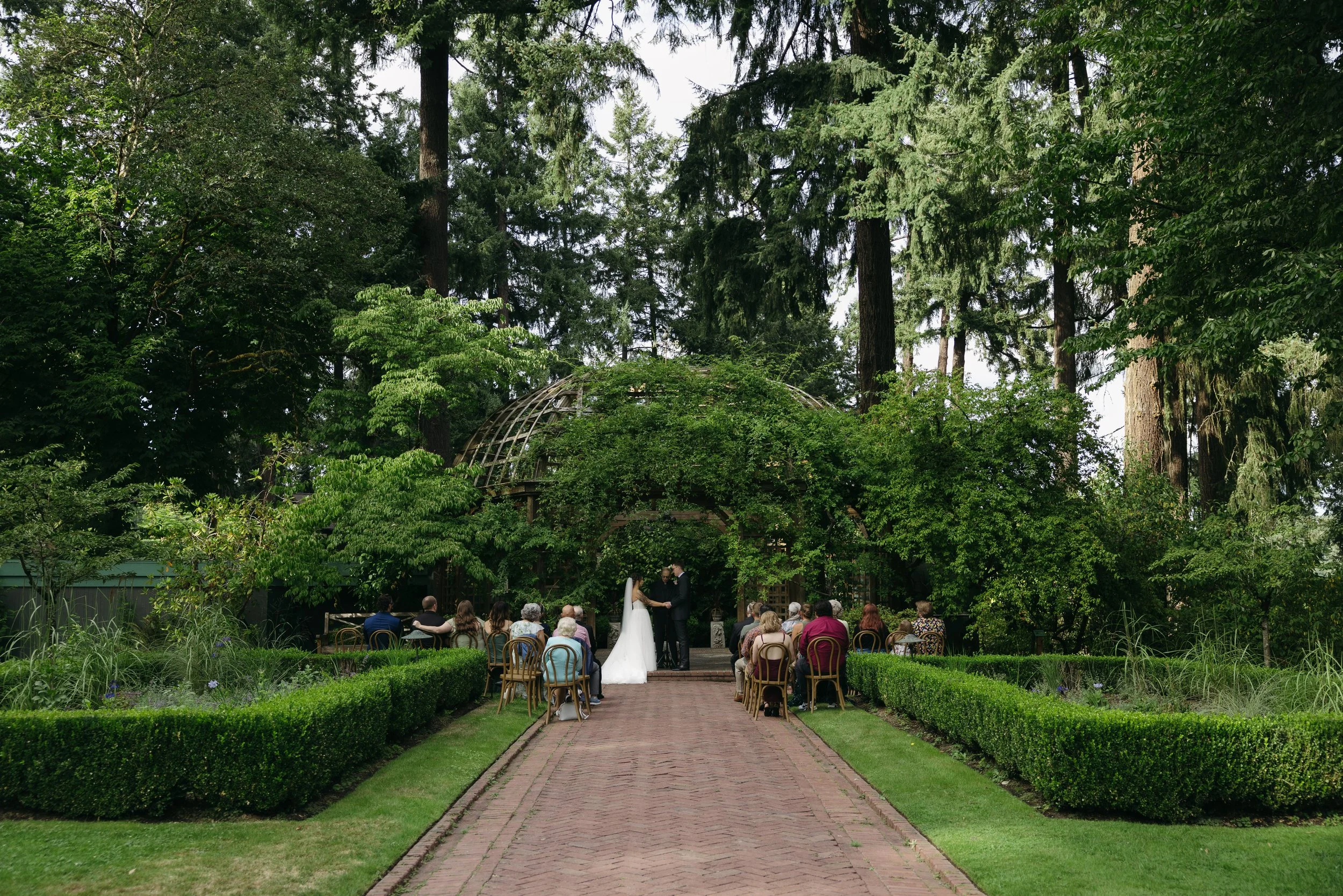 A wedding ceremony taking place outdoors on a garden pathway, with the bride and groom standing under a lush, green arch, surrounded by seated guests, tall trees, and greenery at a European inspired Lakewold Gardens wedding in Seattle