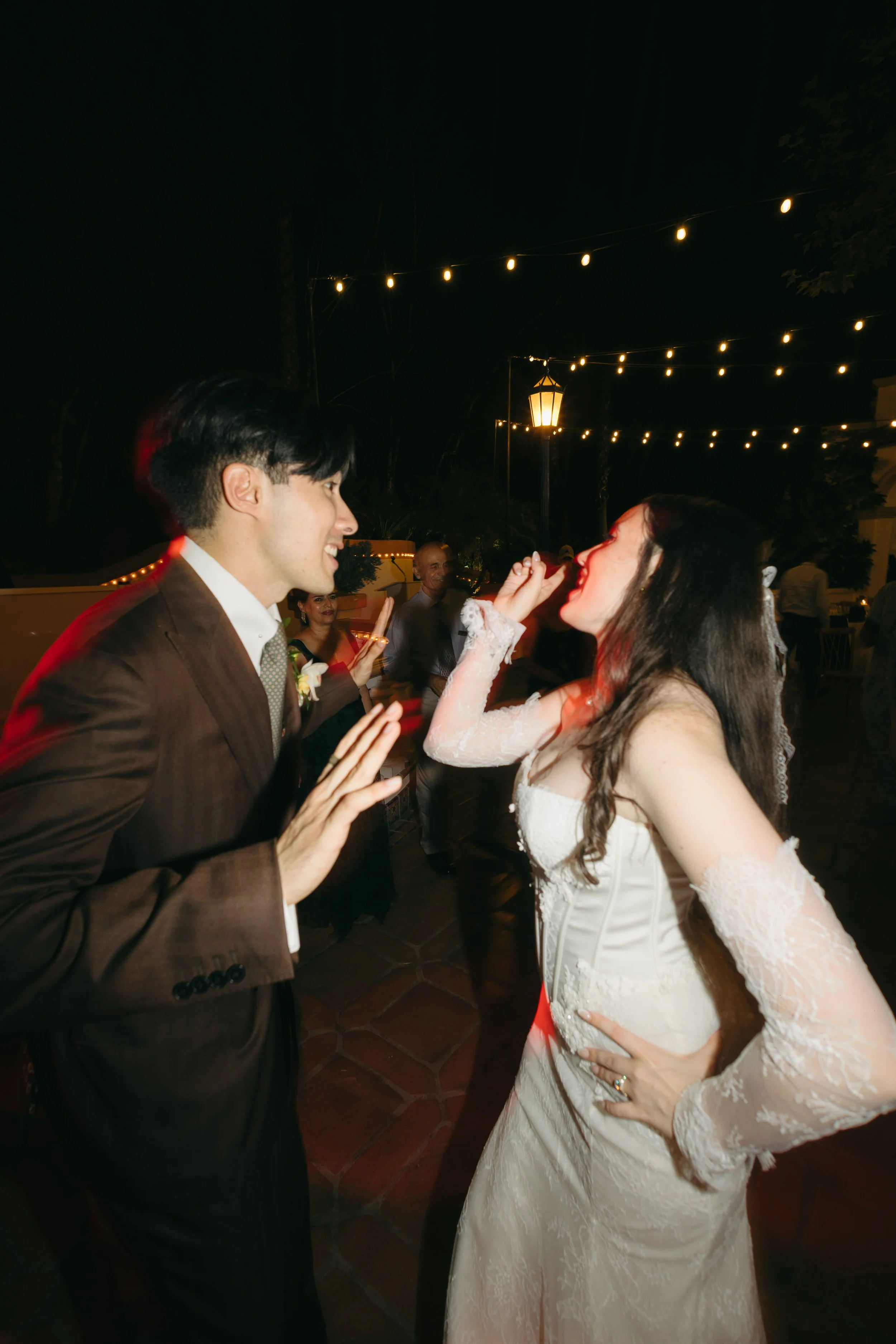 A bride and groom at night, dancing and smiling at each other, with wedding guests in the background under string lights.