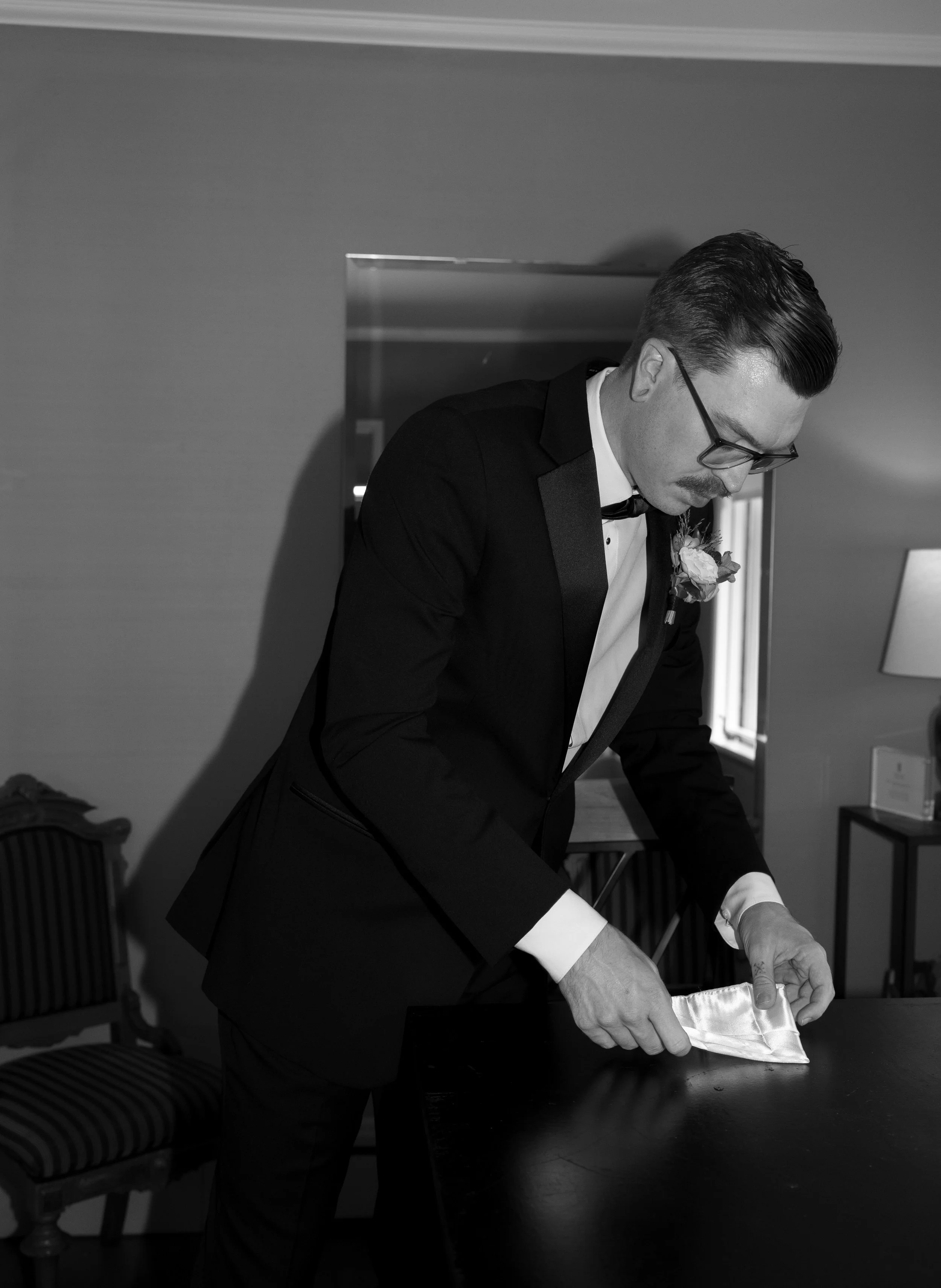 A man in a tuxedo with glasses is folding a satin napkin on a table in a room with chairs, a lamp, and a window.