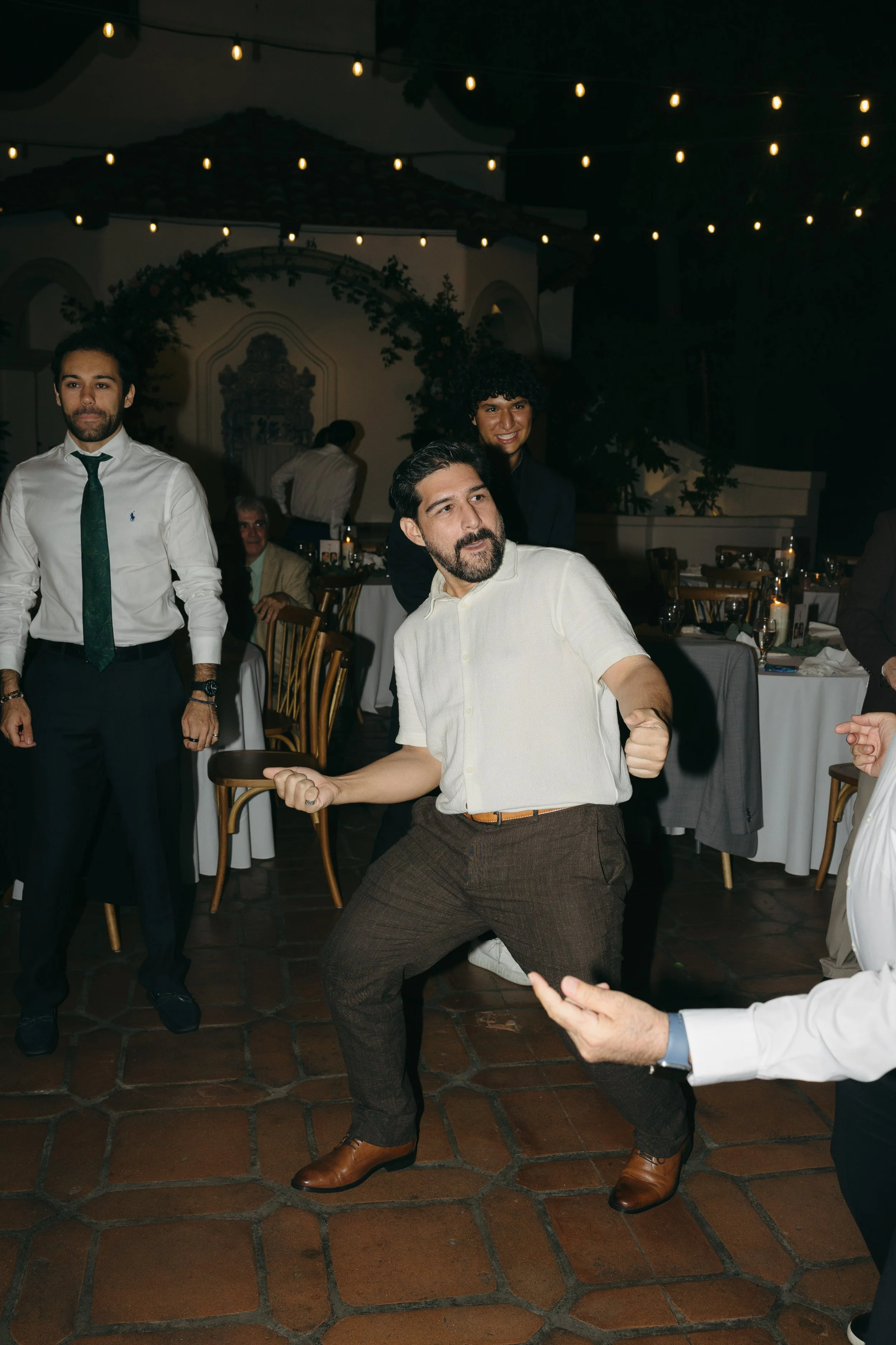 A group of people at an outdoor event dancing and having fun at night, under string lights.