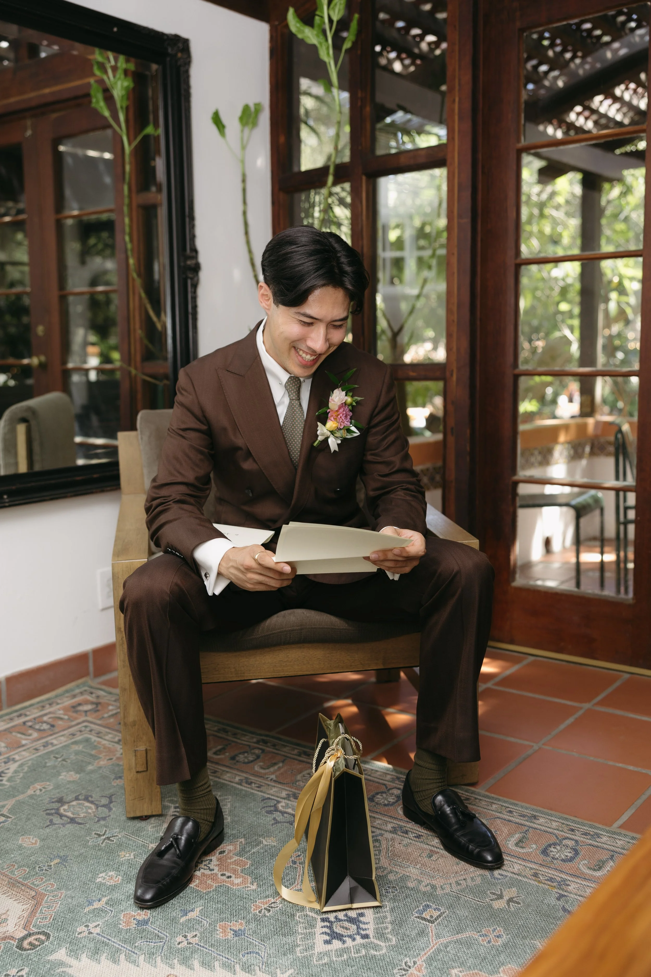 A man in a brown suit with a boutonniere reading a card, sitting on a wooden bench in a sunlit room with wooden framed glass door and windows, a patterned rug, and gifts on the floor.