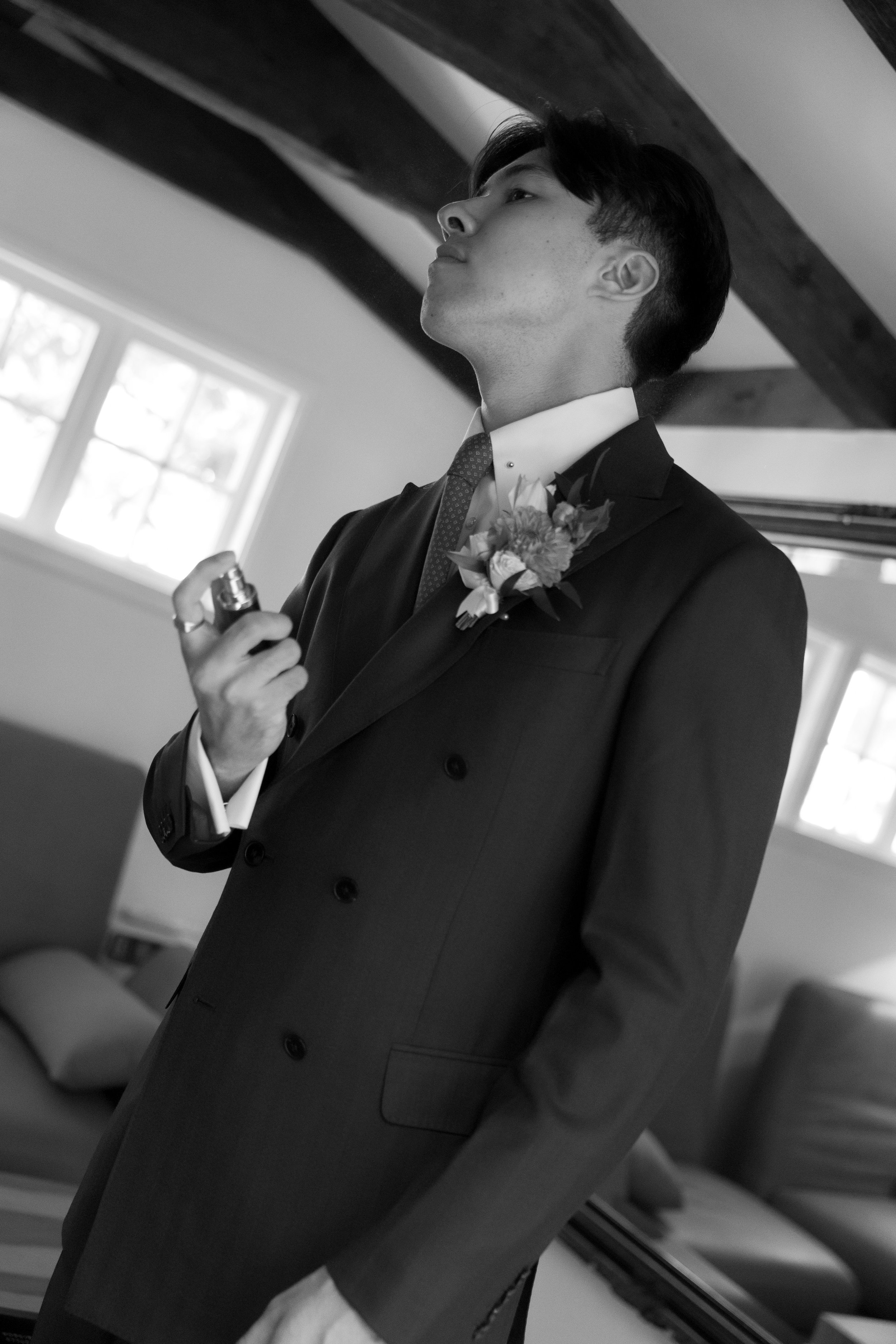 A young man in a tuxedo with a boutonnière, holding a small spray bottle, standing indoors near a mirror and a couch, with wooden beams on the ceiling.