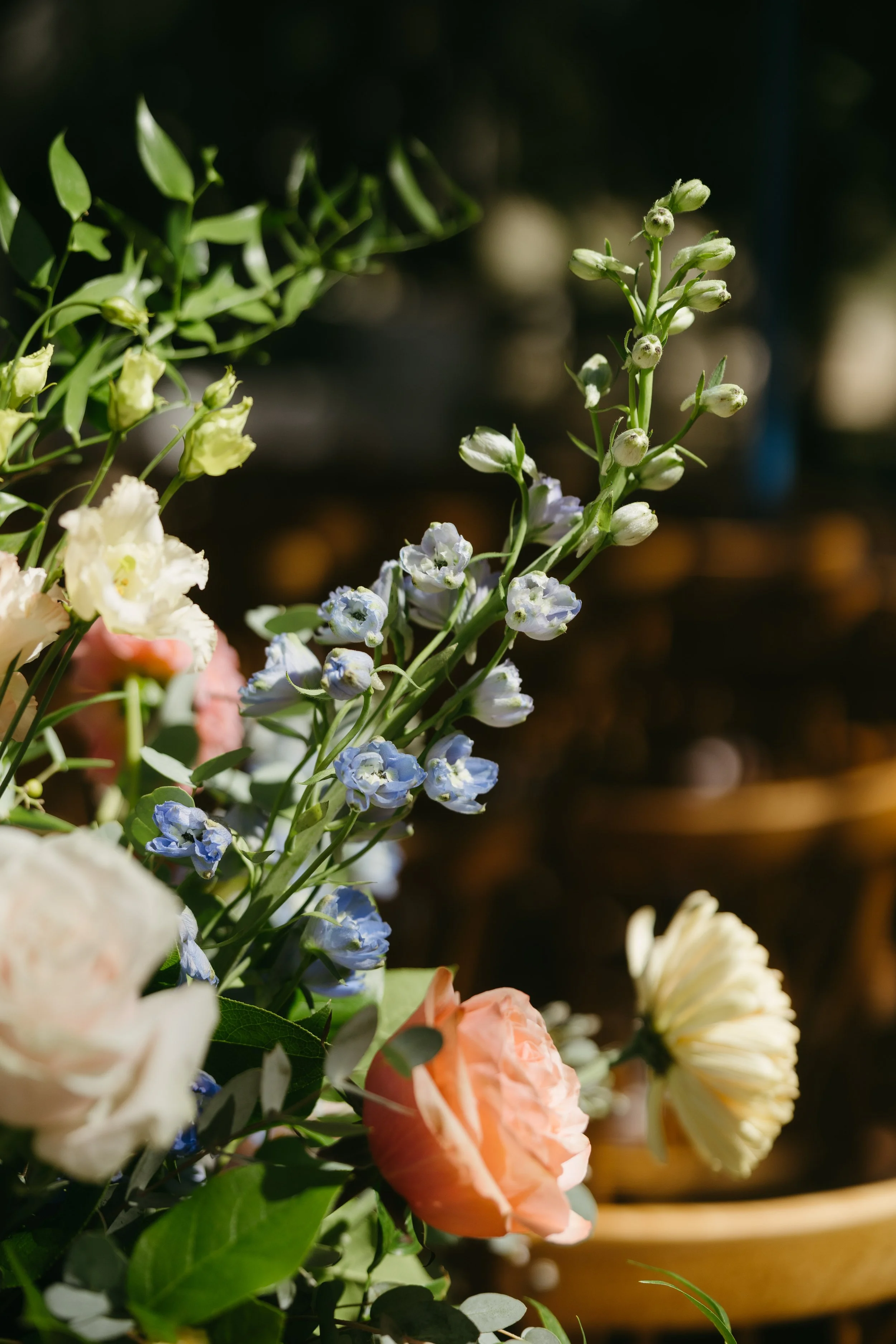 Close-up of a colorful flower arrangement with white, blue, peach, and cream flowers and greenery, set against a dark blurred background.