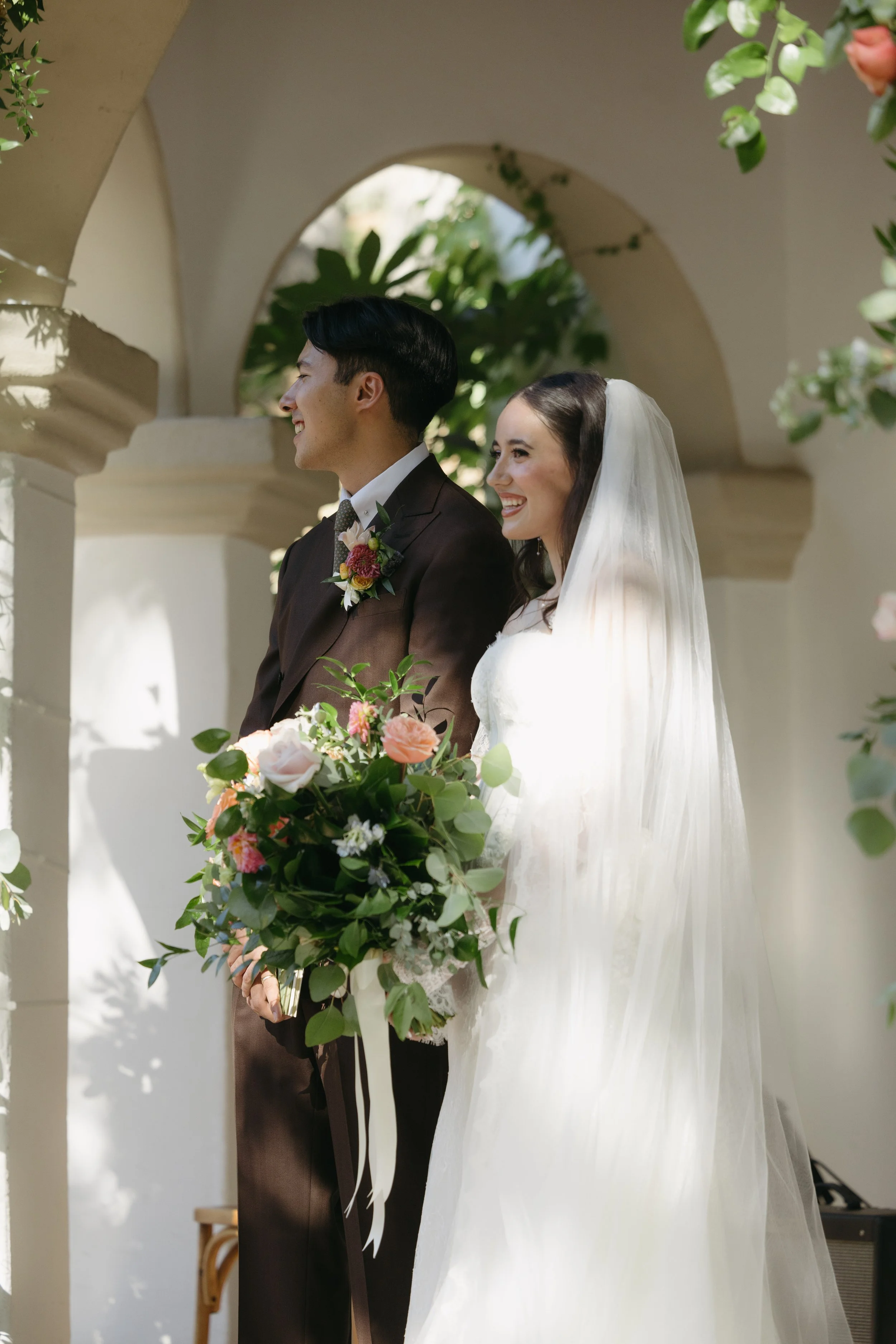 A bride and groom smiling during their wedding ceremony, standing under an archway adorned with greenery and flowers, with the bride holding a bouquet of flowers.