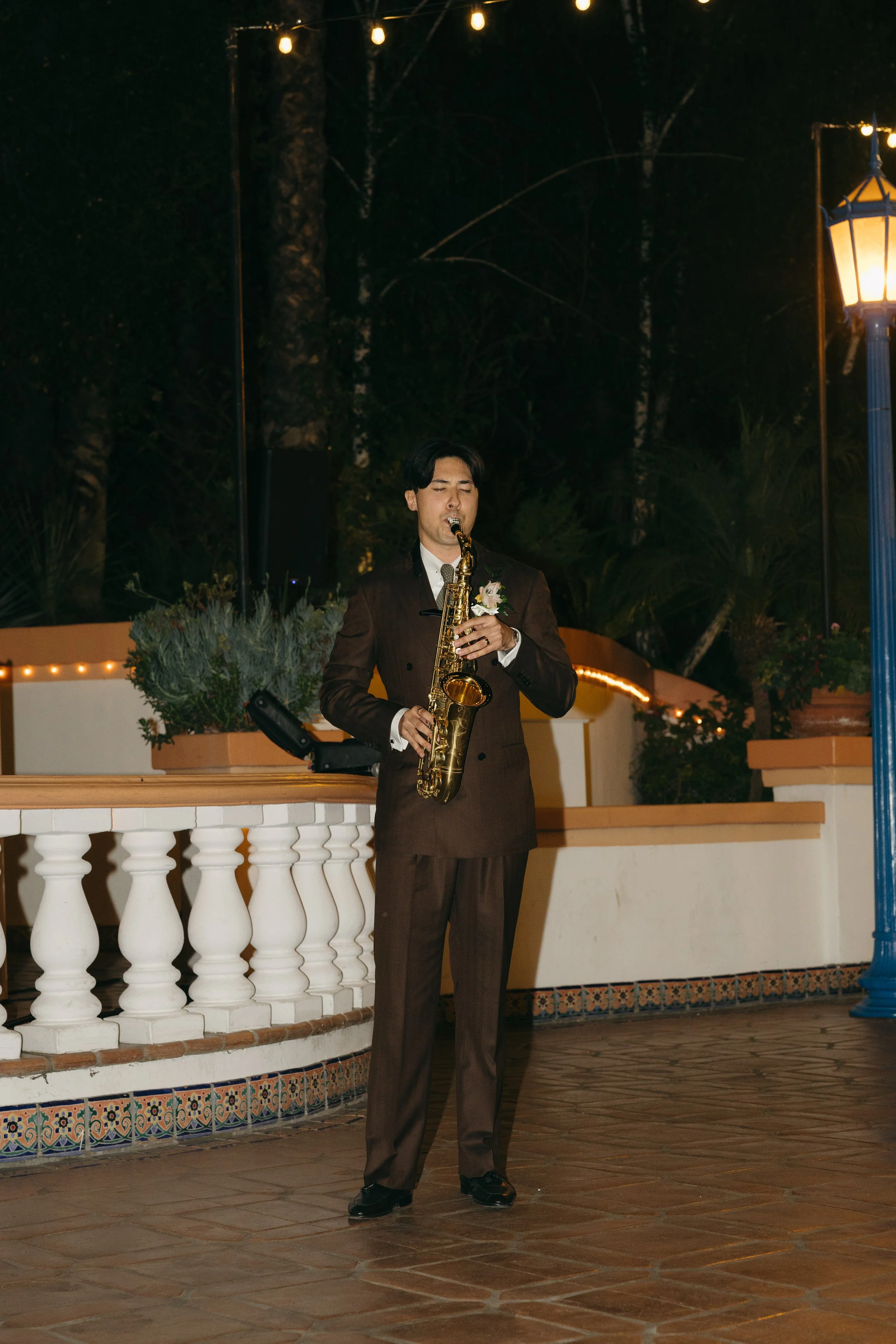 A man in a formal brown suit playing a saxophone at night outdoors, decorated with string lights and surrounded by trees.