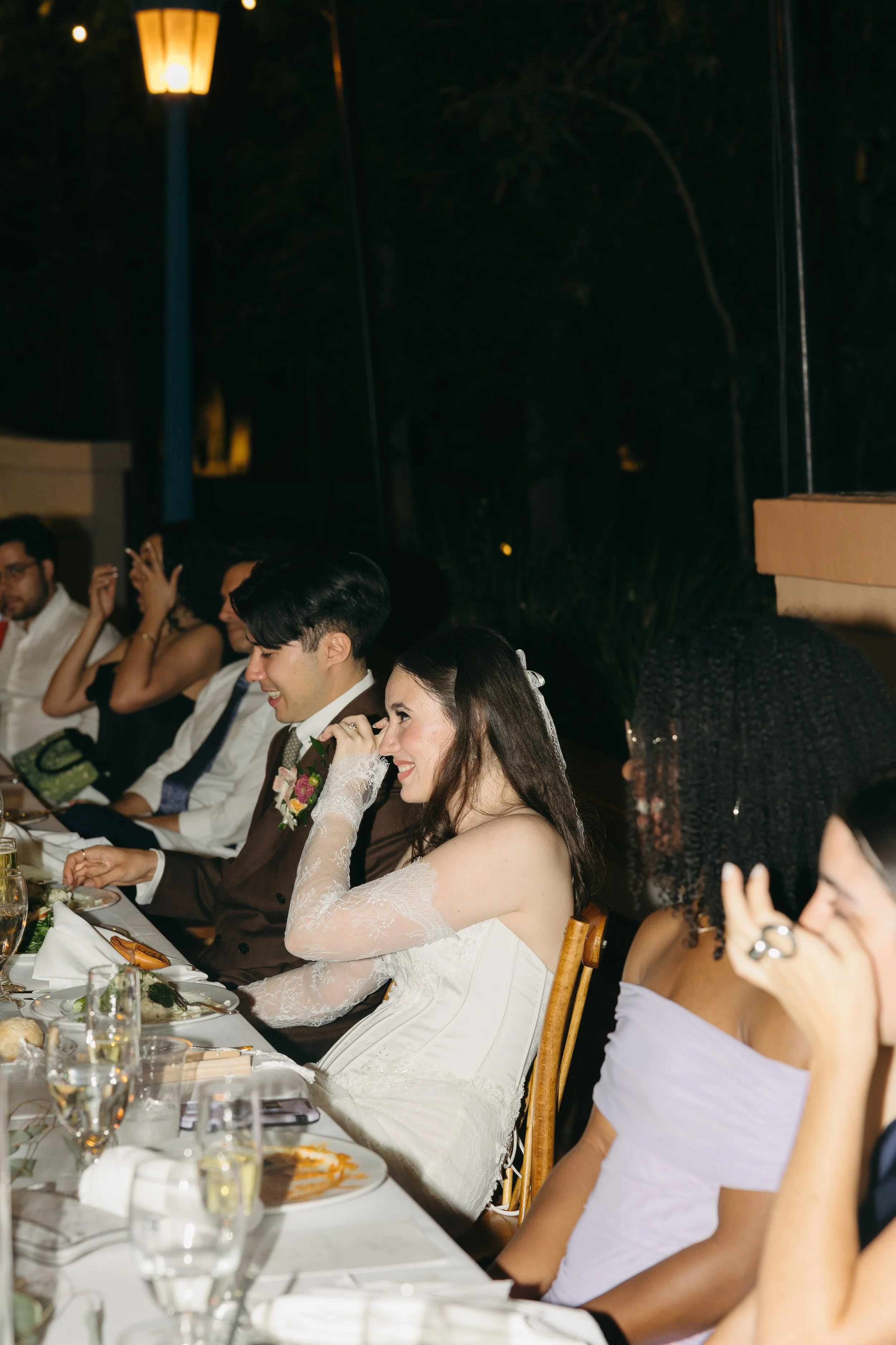People sitting at a long dinner table outdoors during a wedding reception. The bride is wearing a white wedding dress with lace sleeves, and is smiling while touching her face. The groom is wearing a dark suit with a boutonniere, and is smiling. The 