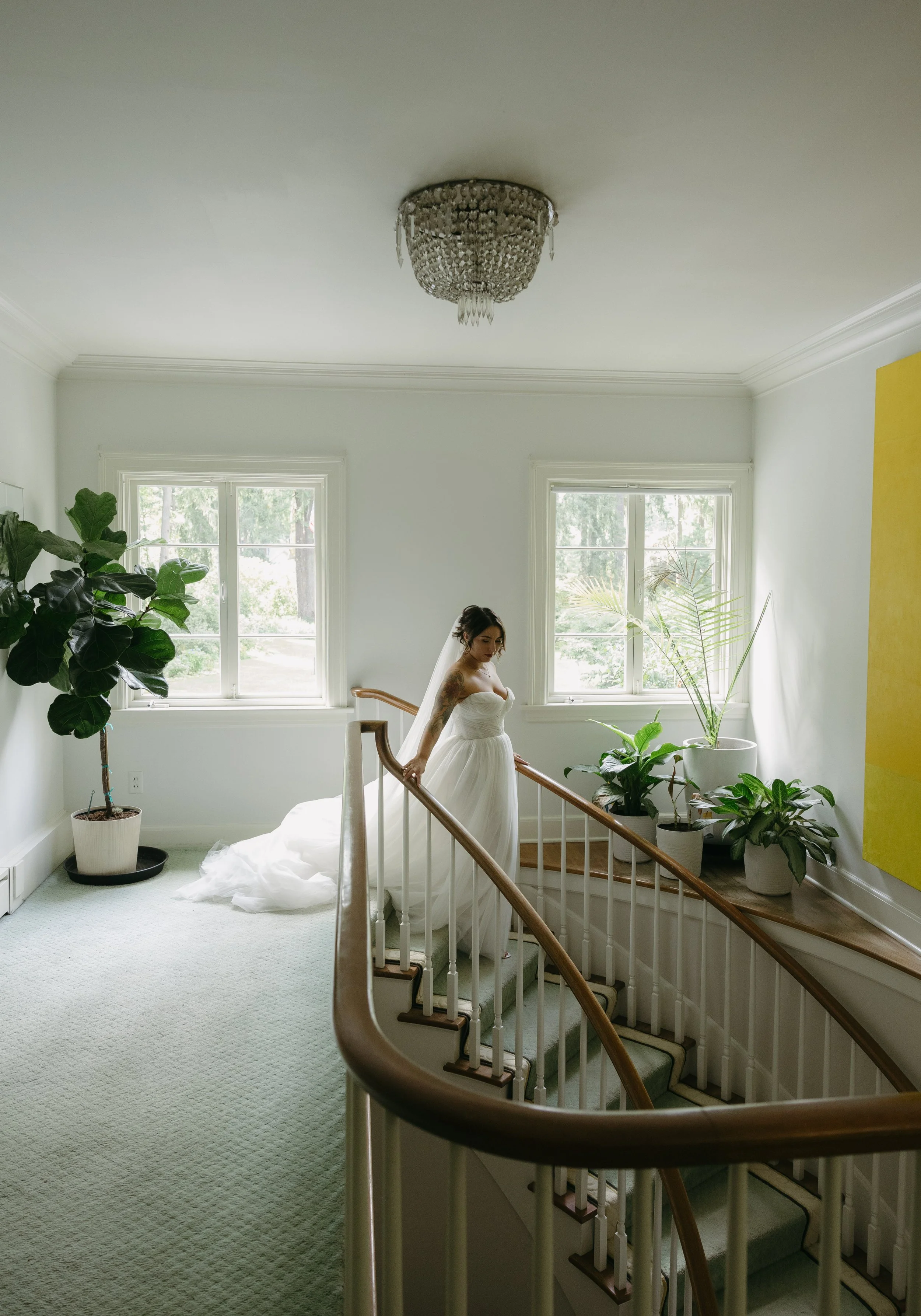 A bride in a white wedding gown standing on a staircase landing, holding the handrail, with large green potted plants nearby, in a bright room with two windows at a European inspired Lakewold Gardens wedding in Seattle