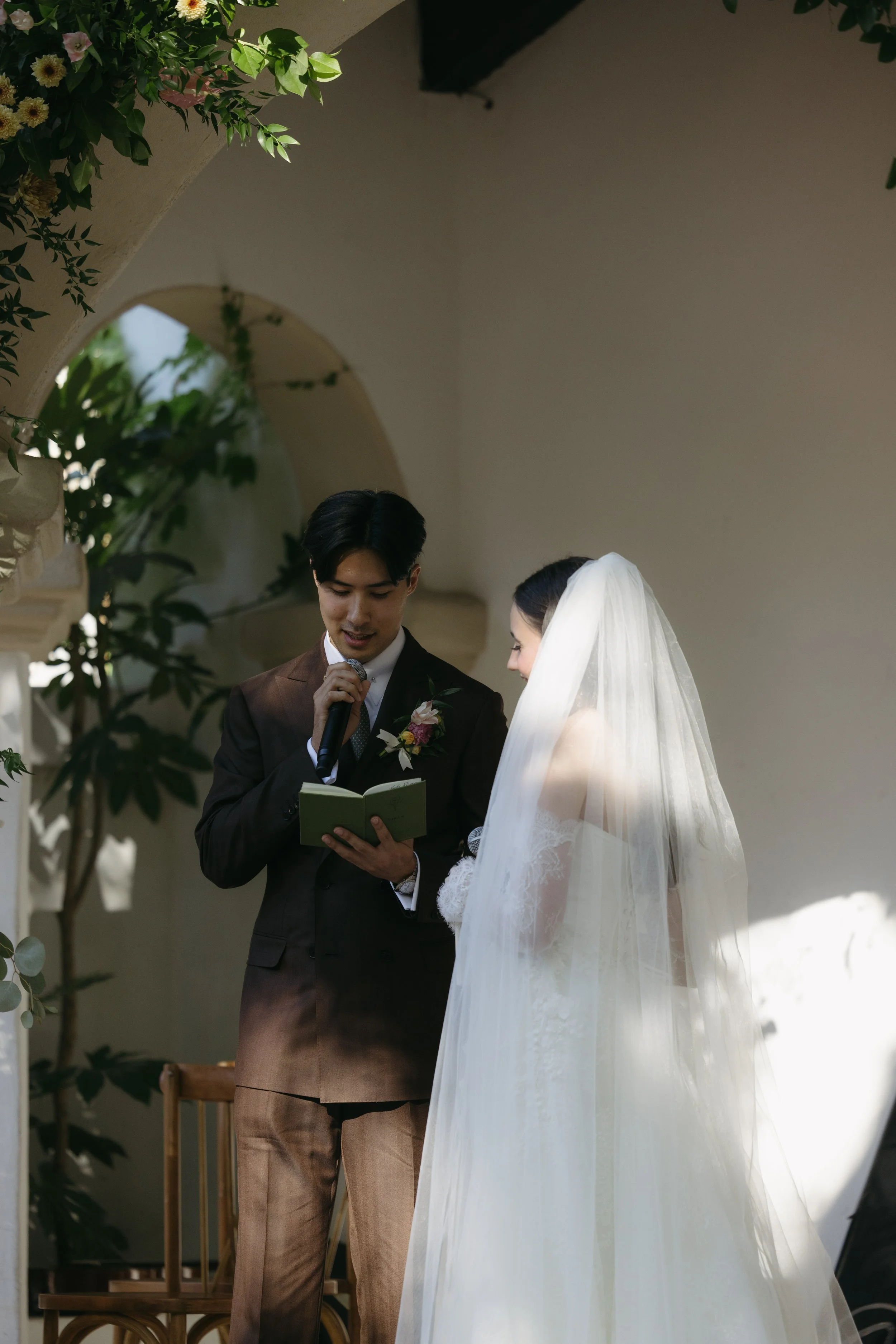 A bride and groom standing outdoors during their wedding ceremony, the groom reading vows from a small book and holding a microphone, with greenery and arches in the background.