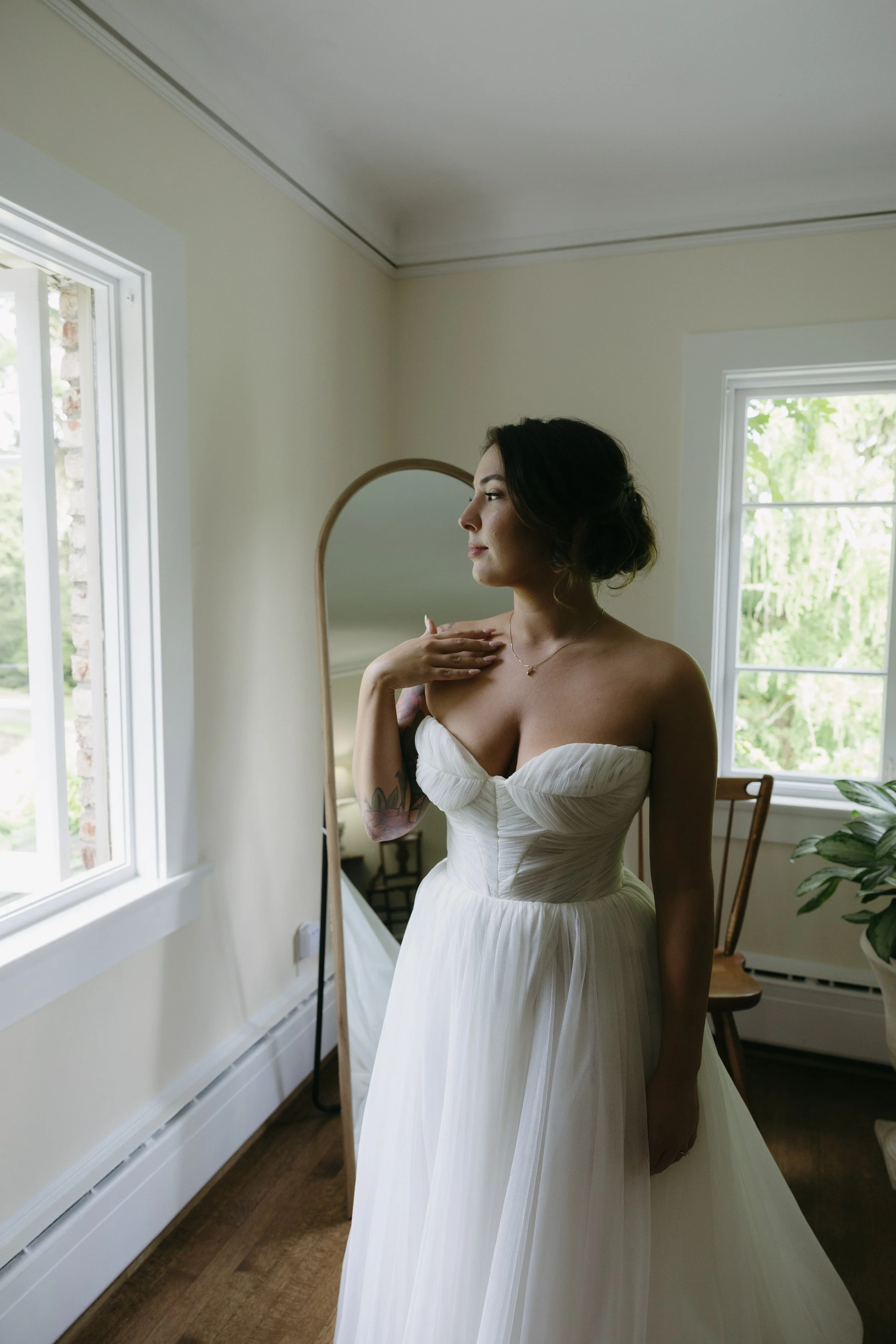 A woman in a white wedding dress standing near a mirror in a bright room with windows, greenery outside, and a wooden chair.