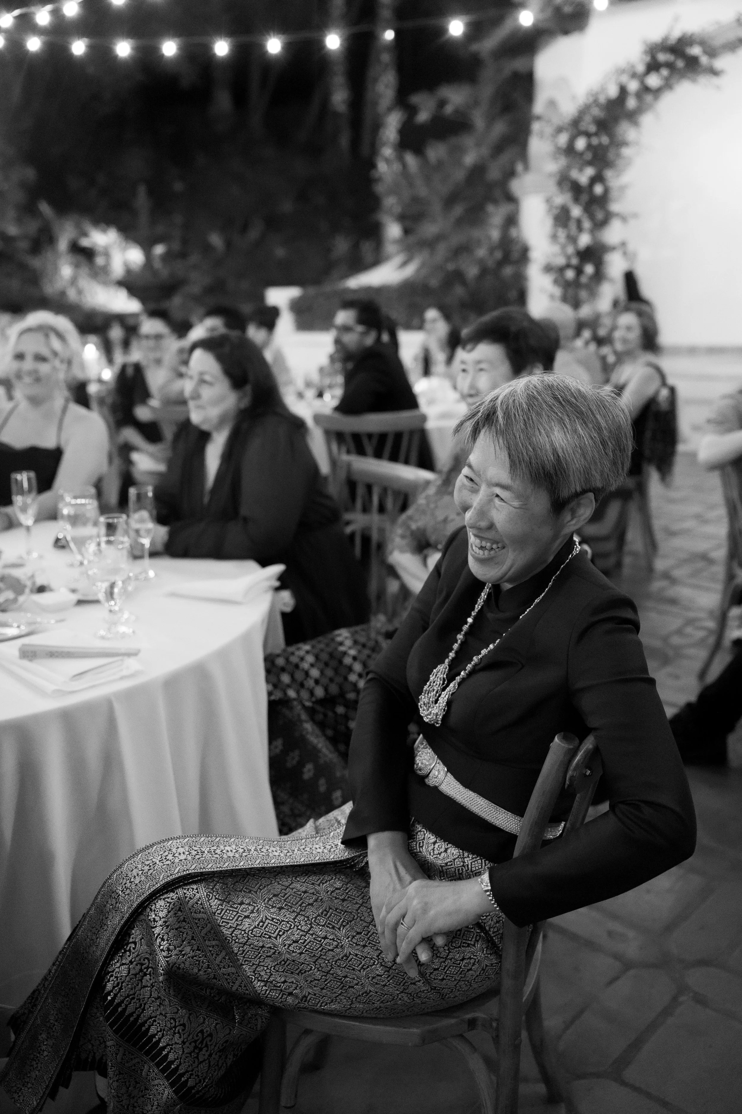A woman in traditional attire smiling and laughing at a formal dinner event, seated at a table with other guests. The setting appears to be outdoors or in a decorated venue, with string lights overhead.