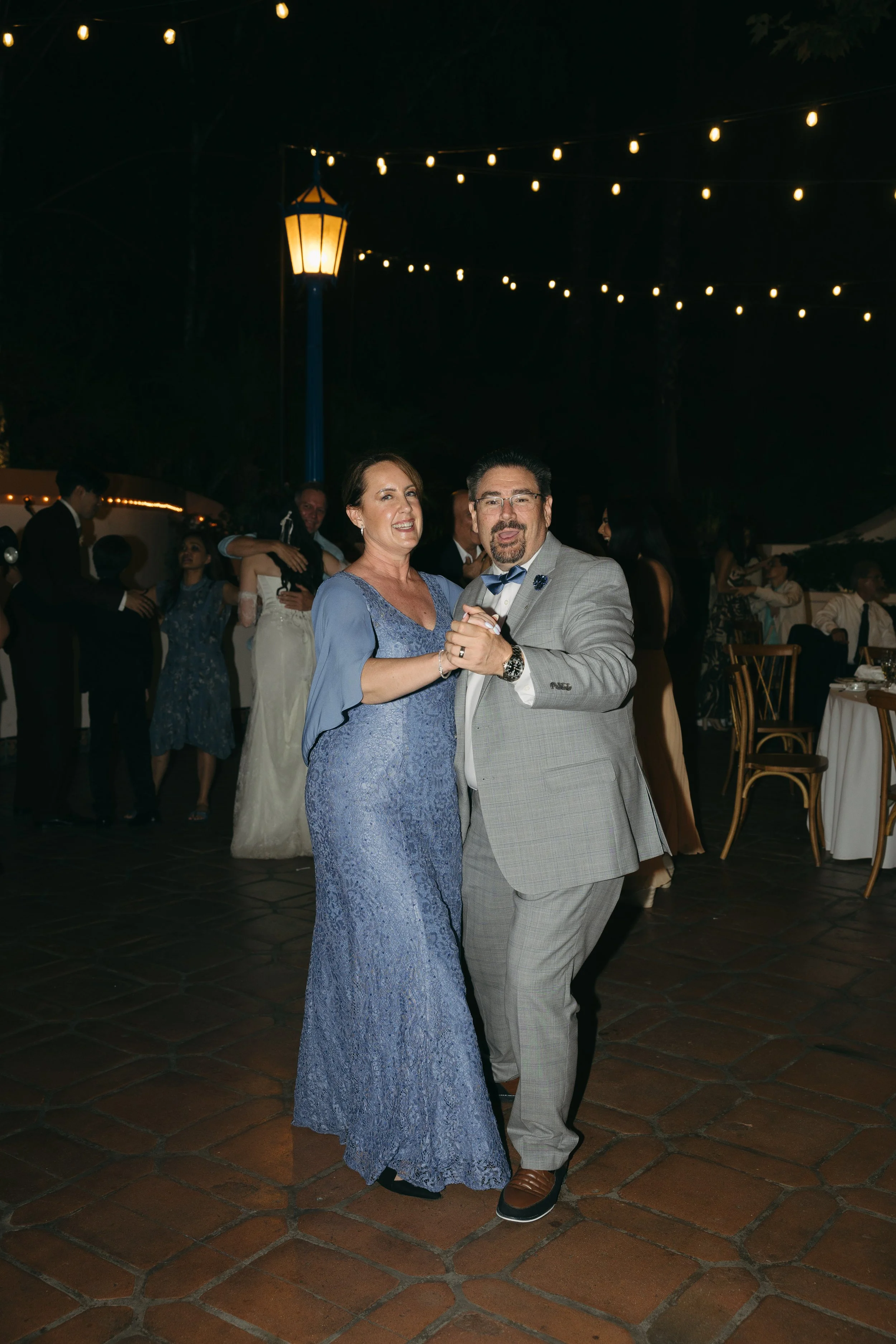 A couple is dancing at a nighttime event, with strings of lights overhead and other people in the background.