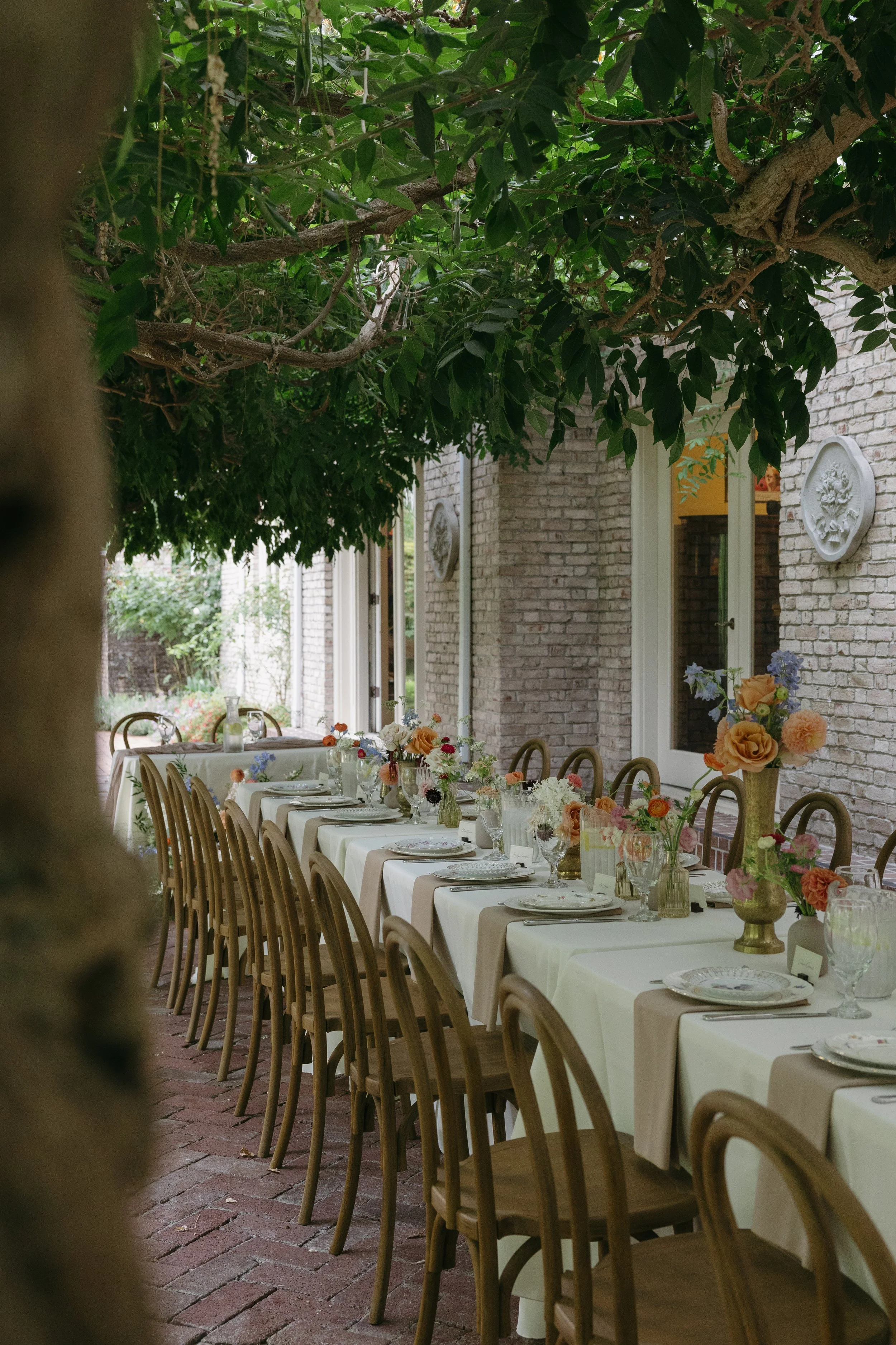 An outdoor reception dinner with white tablecloths, floral centerpieces, plates, and glassware, under the shade of lush green trees on a patio with brick flooring and brick wall in the background at a European inspired Lakewold Gardens wedding in Sea