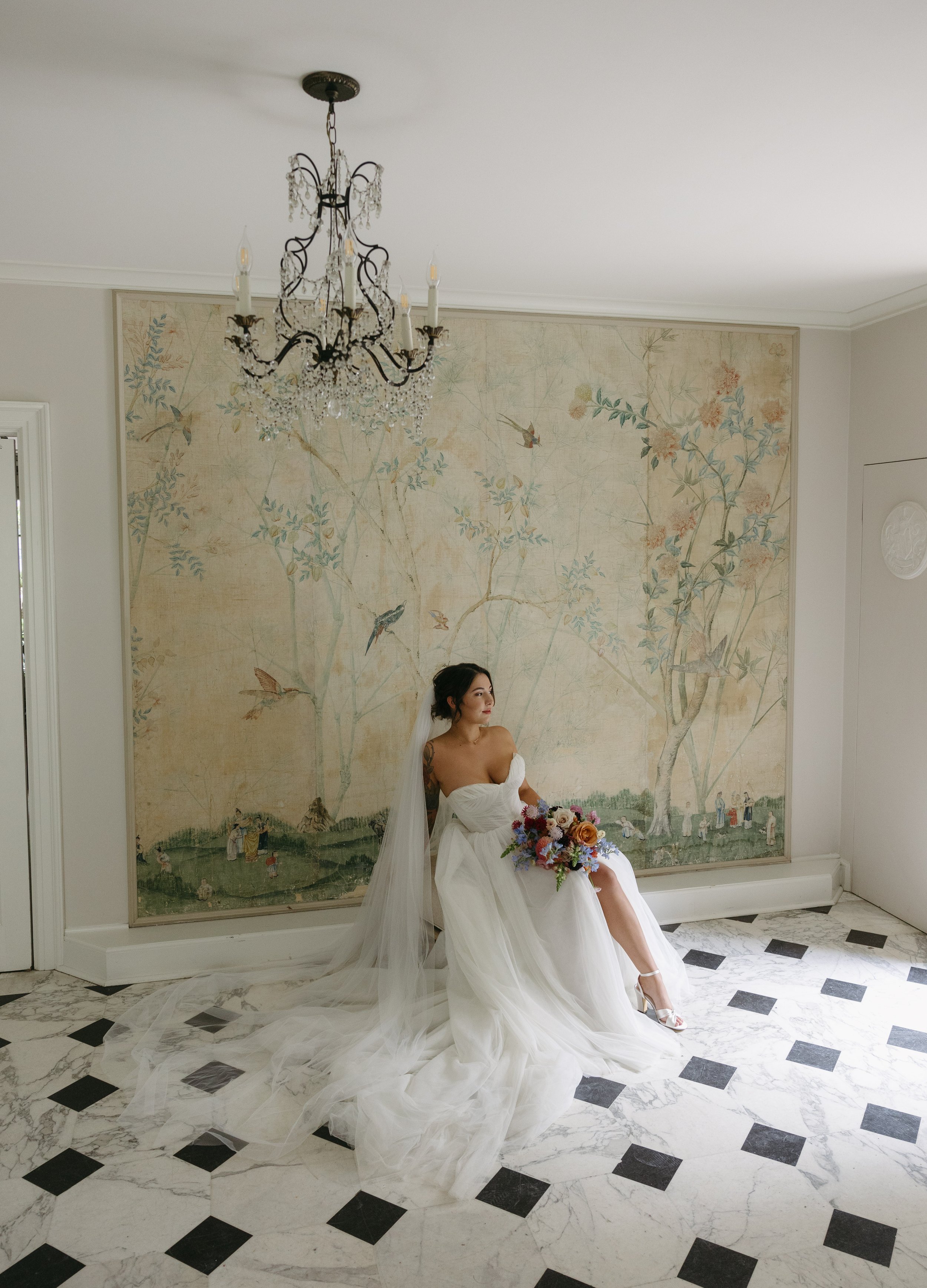 A bride in a white wedding dress sitting in front of a large mural with trees and birds, holding a colorful bouquet, with a chandelier overhead and black-and-white checkered marble floor at a European inspired Lakewold Gardens wedding in Seattle