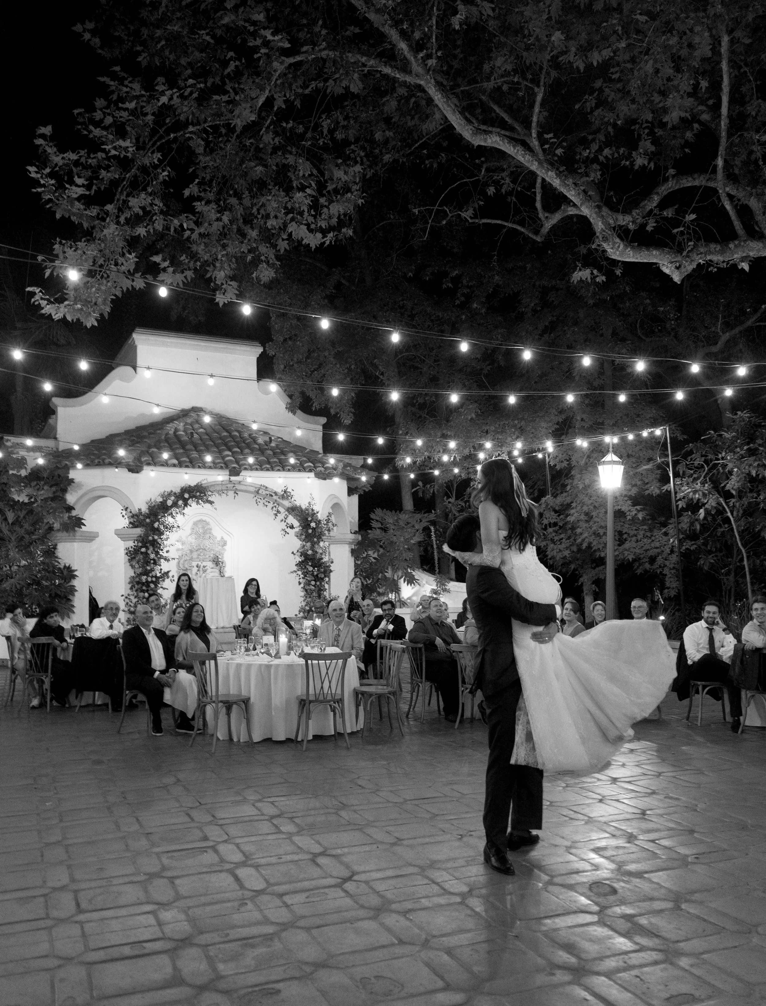 A black and white photo of a wedding reception outdoors at night with string lights, guests seated at round tables, and a bride and groom sharing a dance in the center.