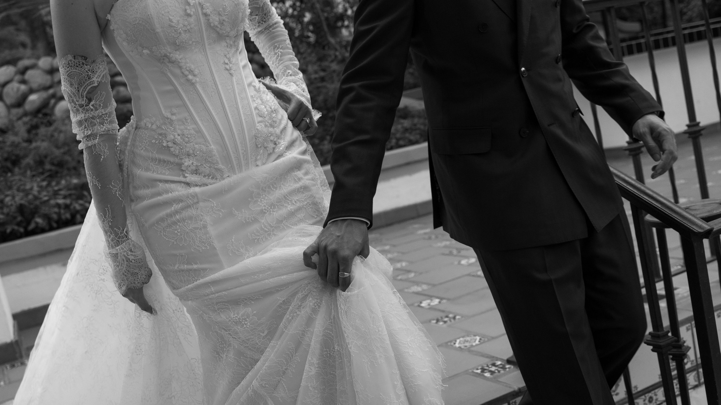 A bride in a wedding dress holding up the hem, and a groom in a suit, walking together on a brick path with flower designs, near a railing.