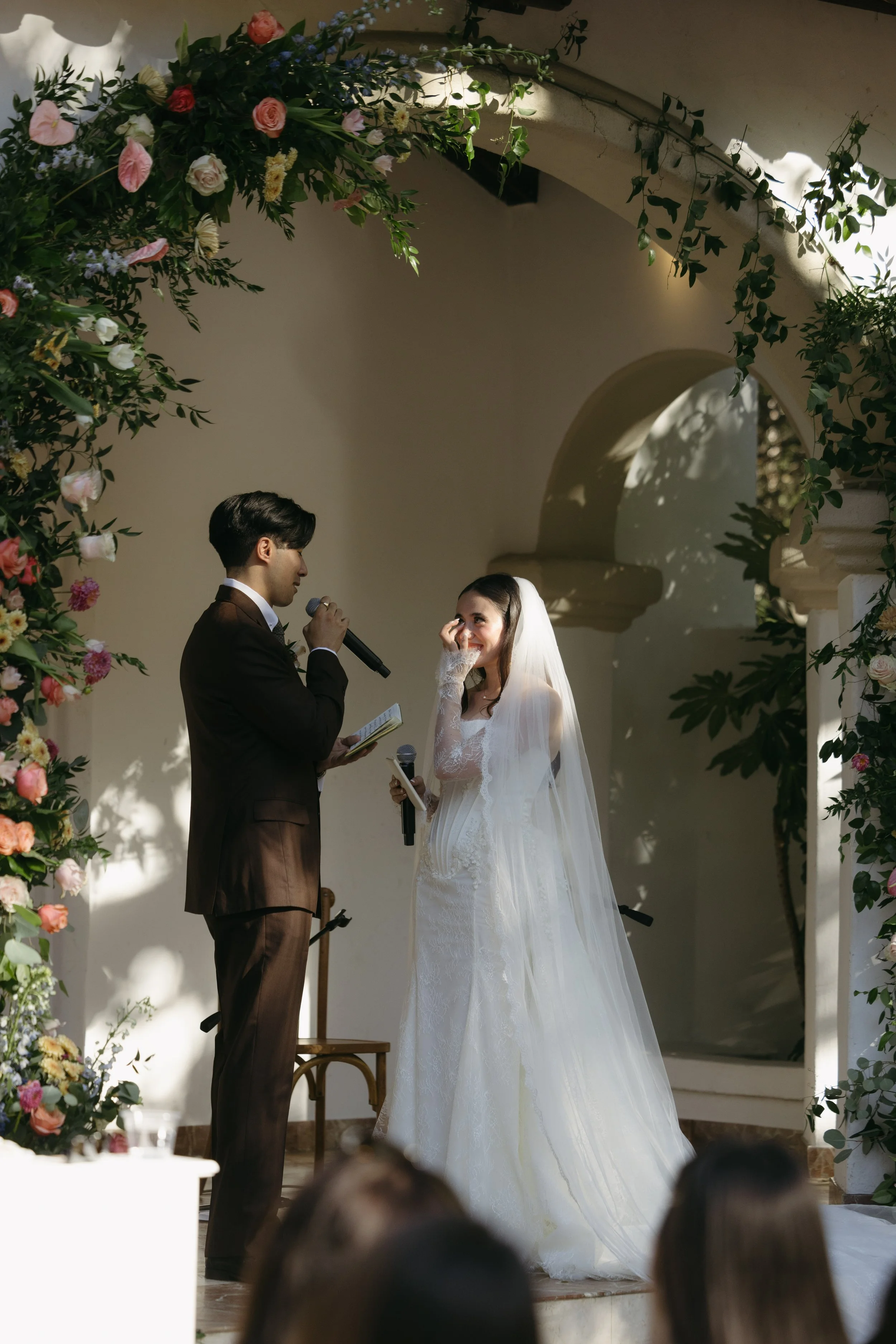A bride and groom exchange vows during their wedding ceremony, framed by floral archways and soft sunlight.