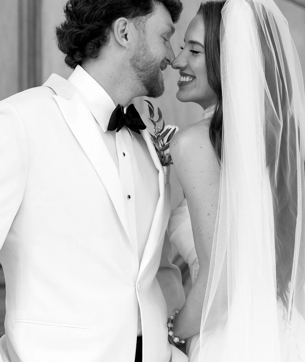 A black and white photo of a wedding couple. The groom, with curly hair and a beard, is in a white tuxedo with a black bow tie. The bride has long hair, a veil, and is smiling with her eyes closed. They are close, touching noses, and appear happy.