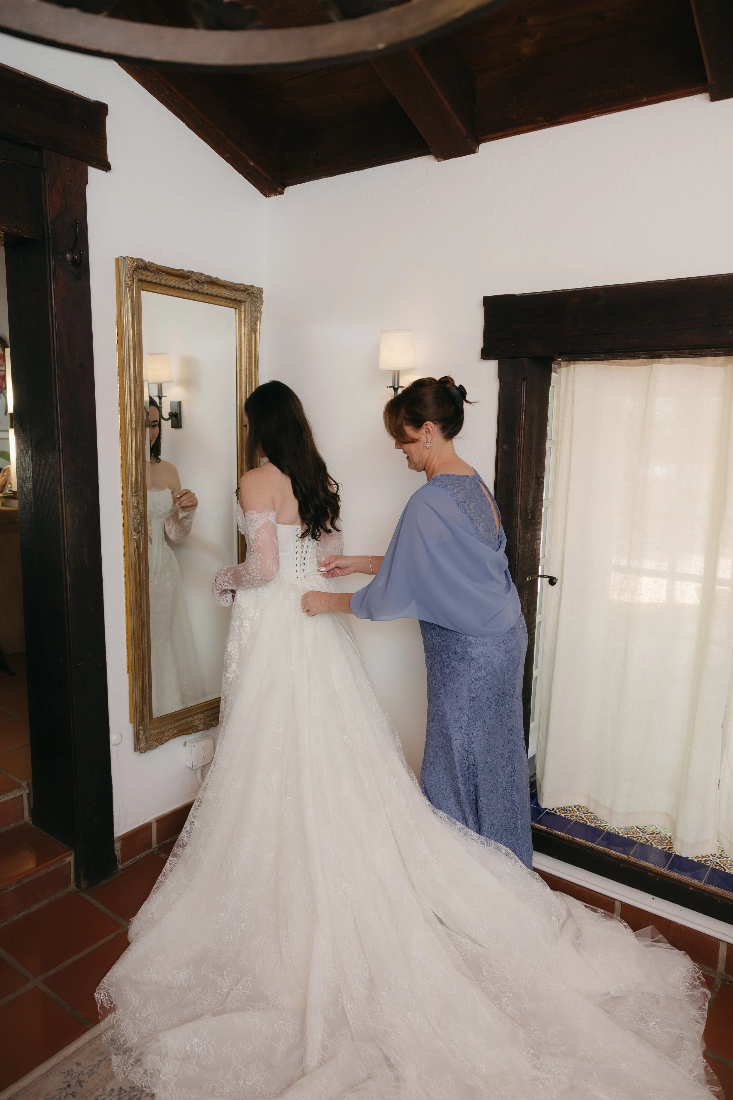 A woman in a wedding dress is being helped by another woman, possibly her mother, to zip up her dress in a room with white walls, dark wood accents, and a large mirror.