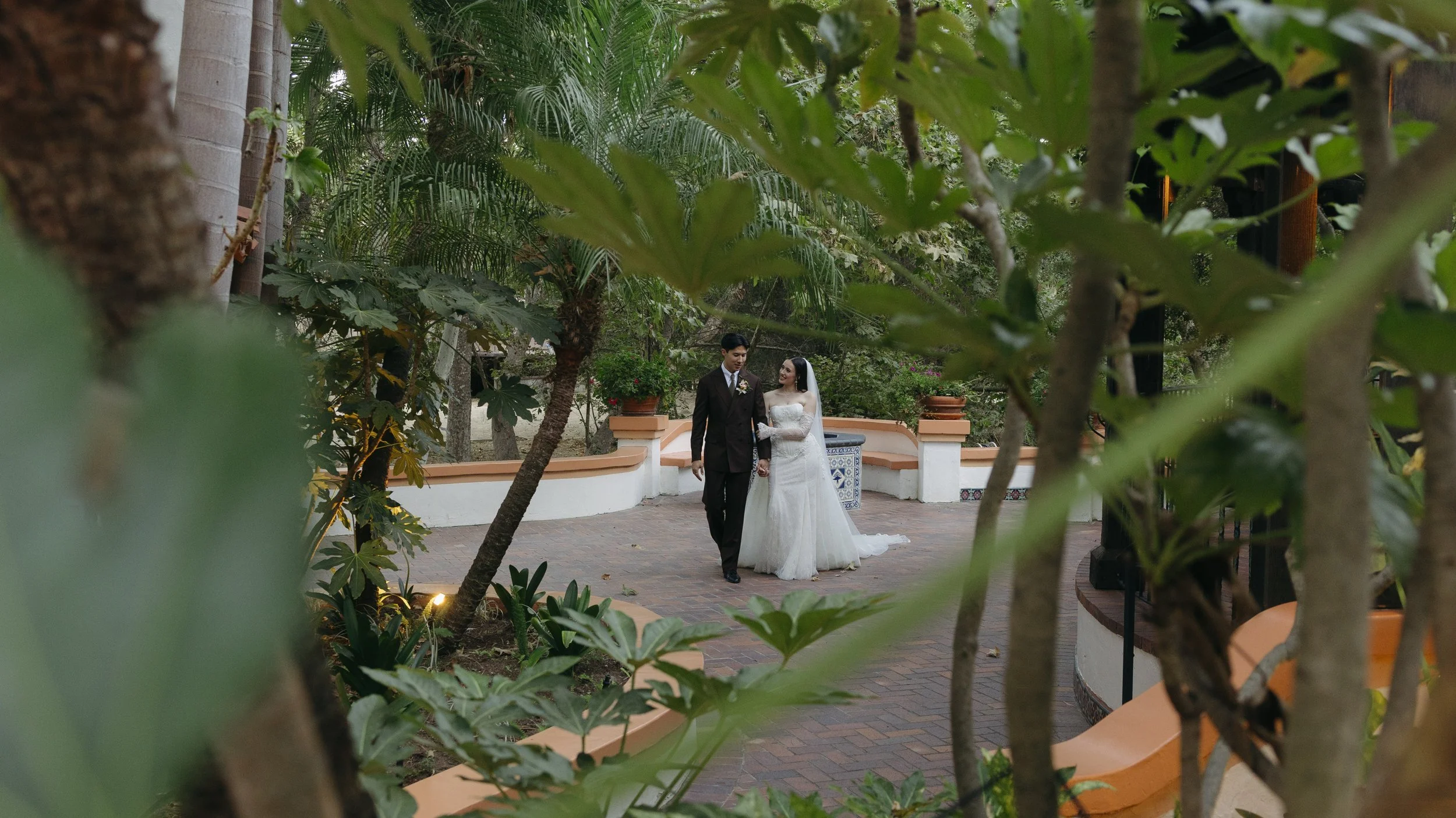 A bride and groom walking hand in hand in an outdoor garden setting, surrounded by lush greenery and potted plants, seen through a frame of tree branches and leaves at a Rancho Las Lomas Wedding in Los Angeles
