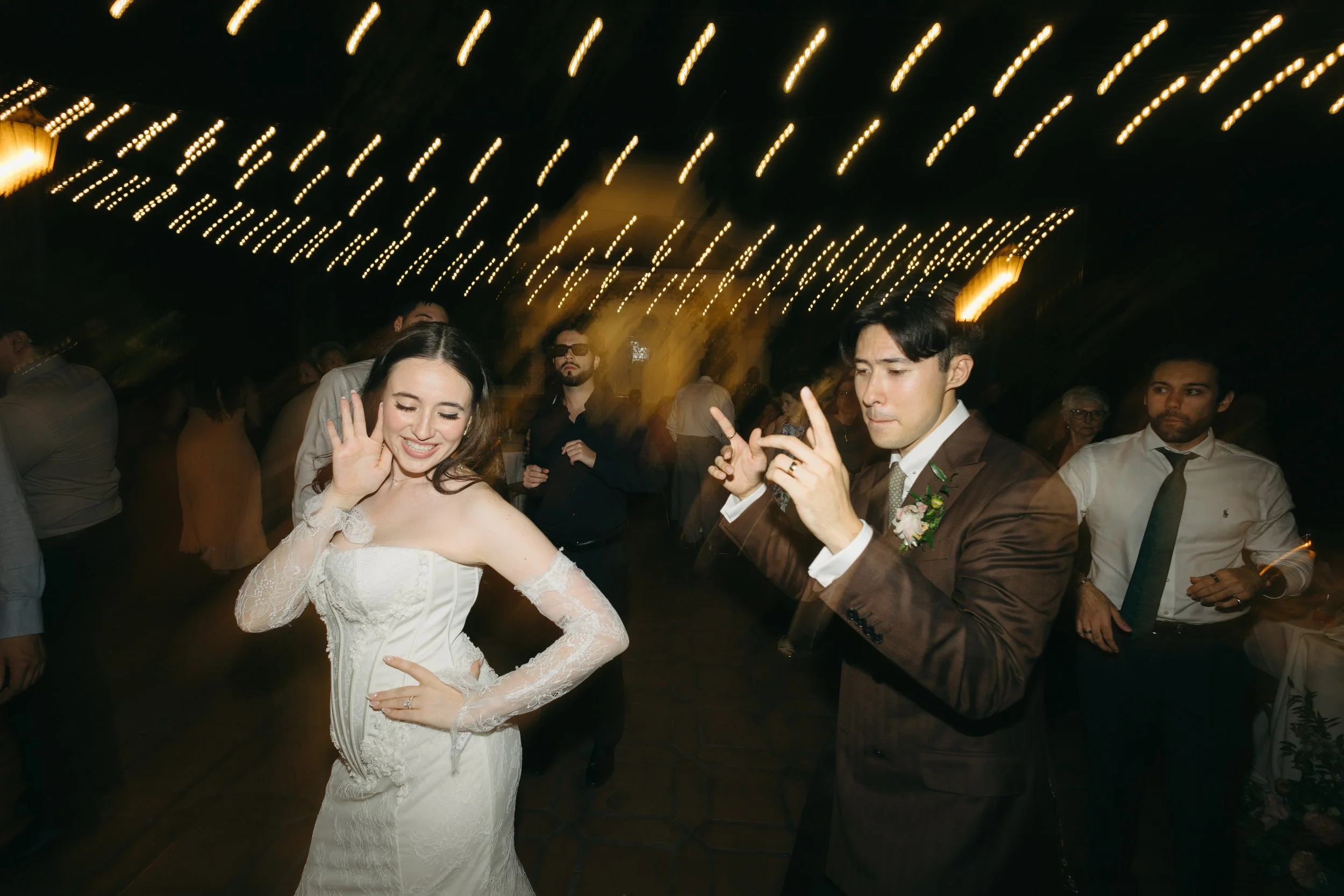 A bride dancing at her wedding reception with a groom and guests under string lights at night.