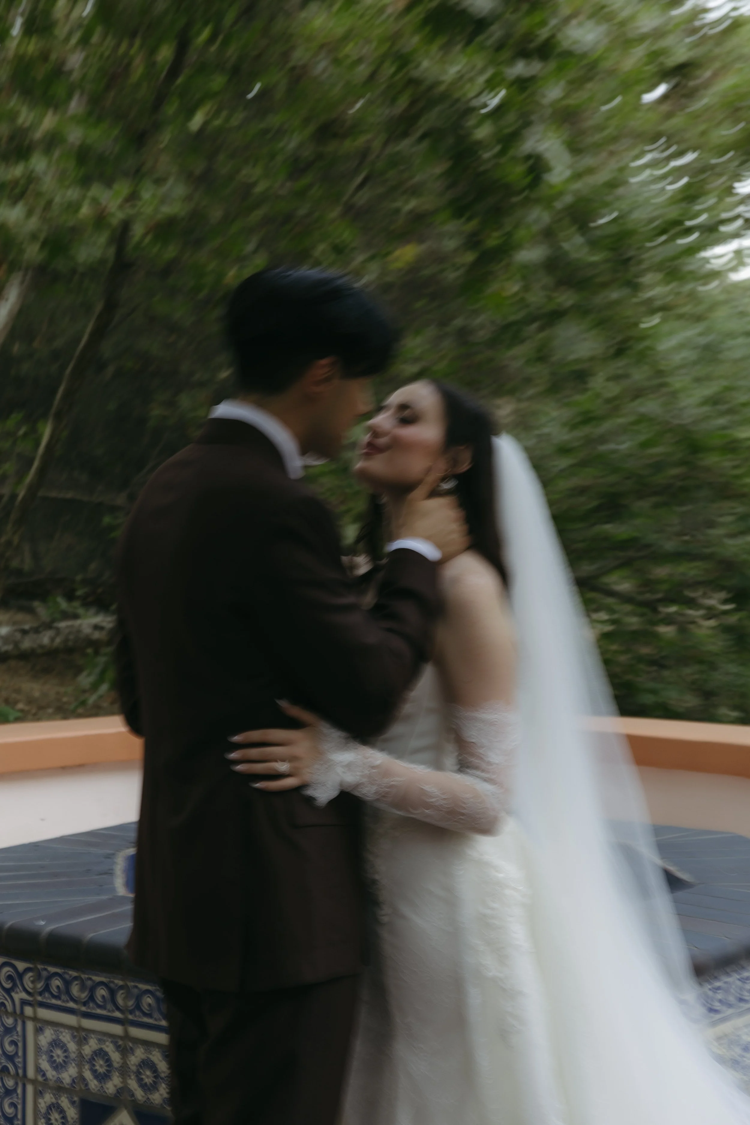 A bride and groom sharing a romantic moment outdoors on their wedding day, with the bride in a white gown and veil and the groom in a suit, standing on a patterned tile surface with green trees in the background.
