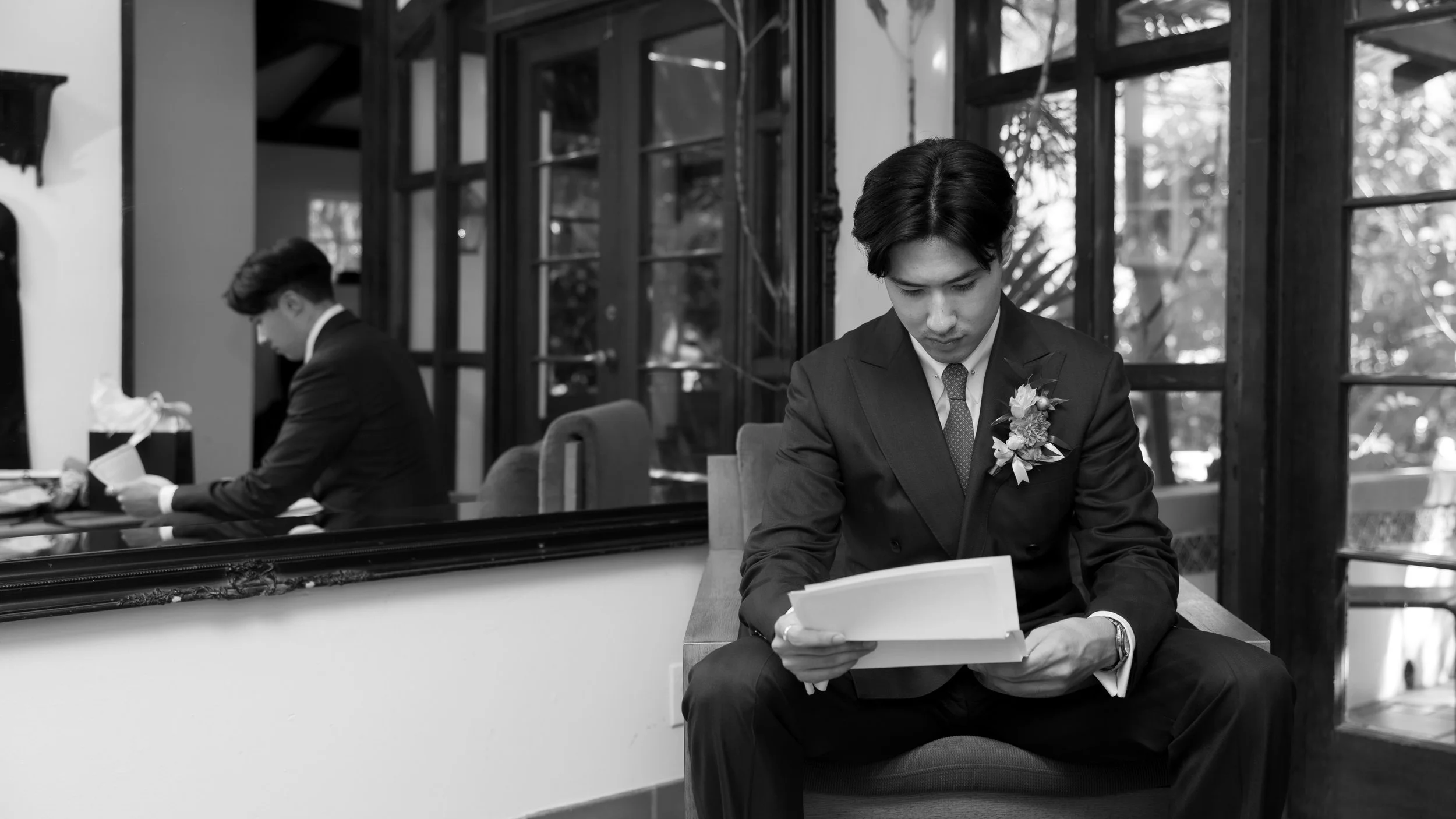 Two young men in formal suits, one sitting and reading papers, the other standing and looking at his phone, in a room with large windows and reflections.