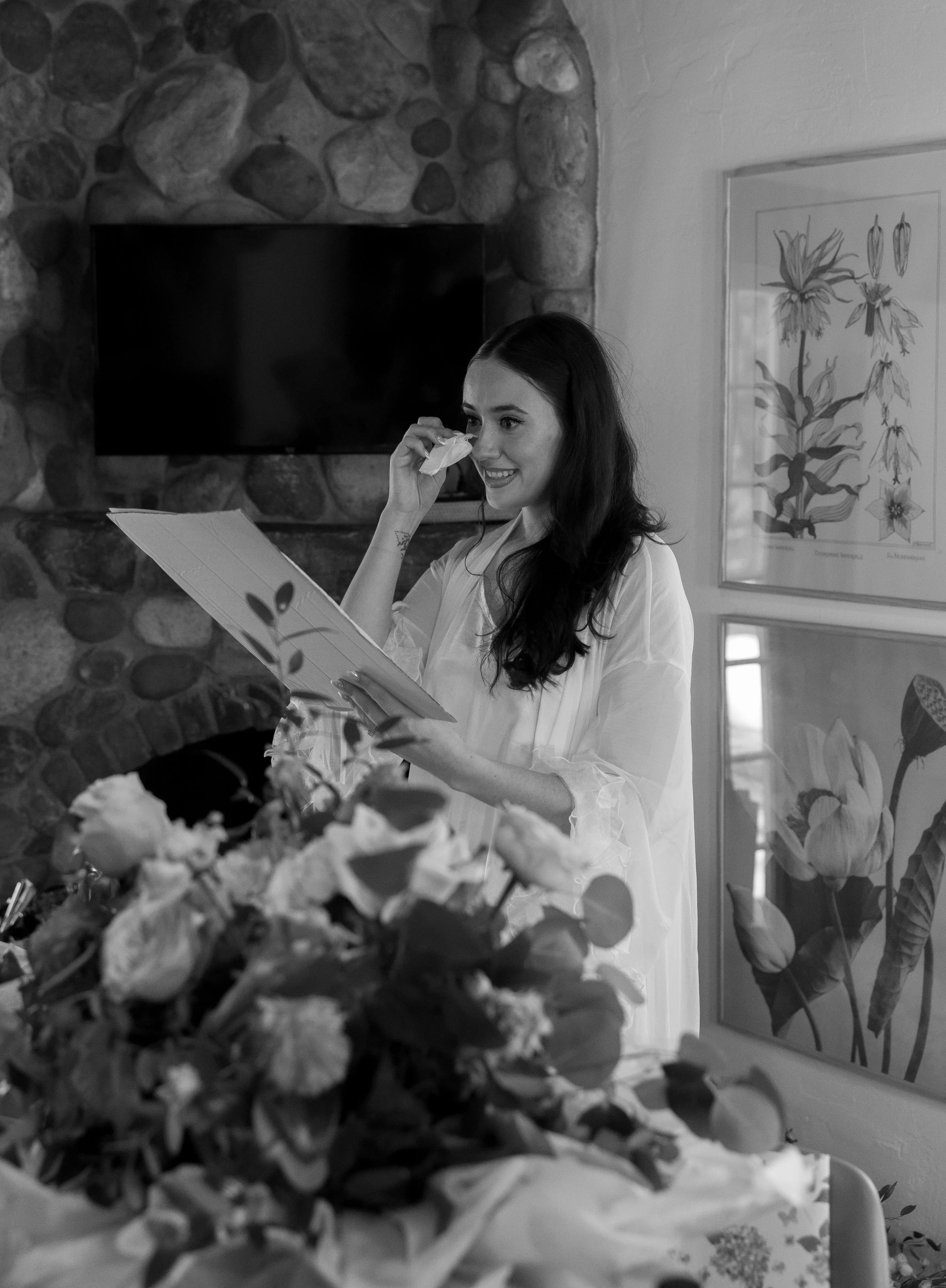 A woman smiling and wiping her nose with a tissue, standing in a room with a stone fireplace and botanical artwork on the wall, surrounded by flowers.