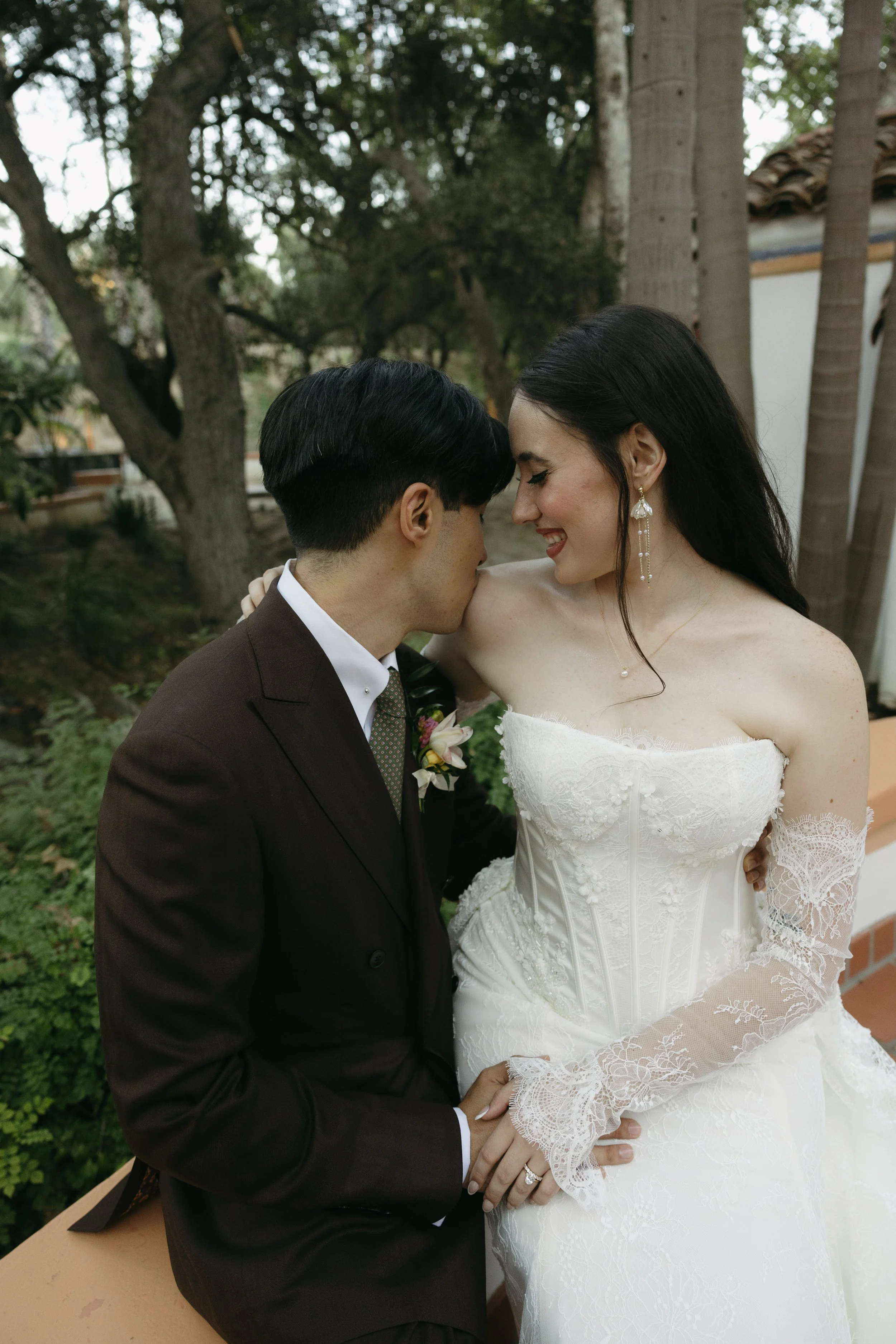 A wedding couple in an outdoor garden, with the bride in a white lace wedding dress and the groom in a dark suit, sharing an intimate moment.