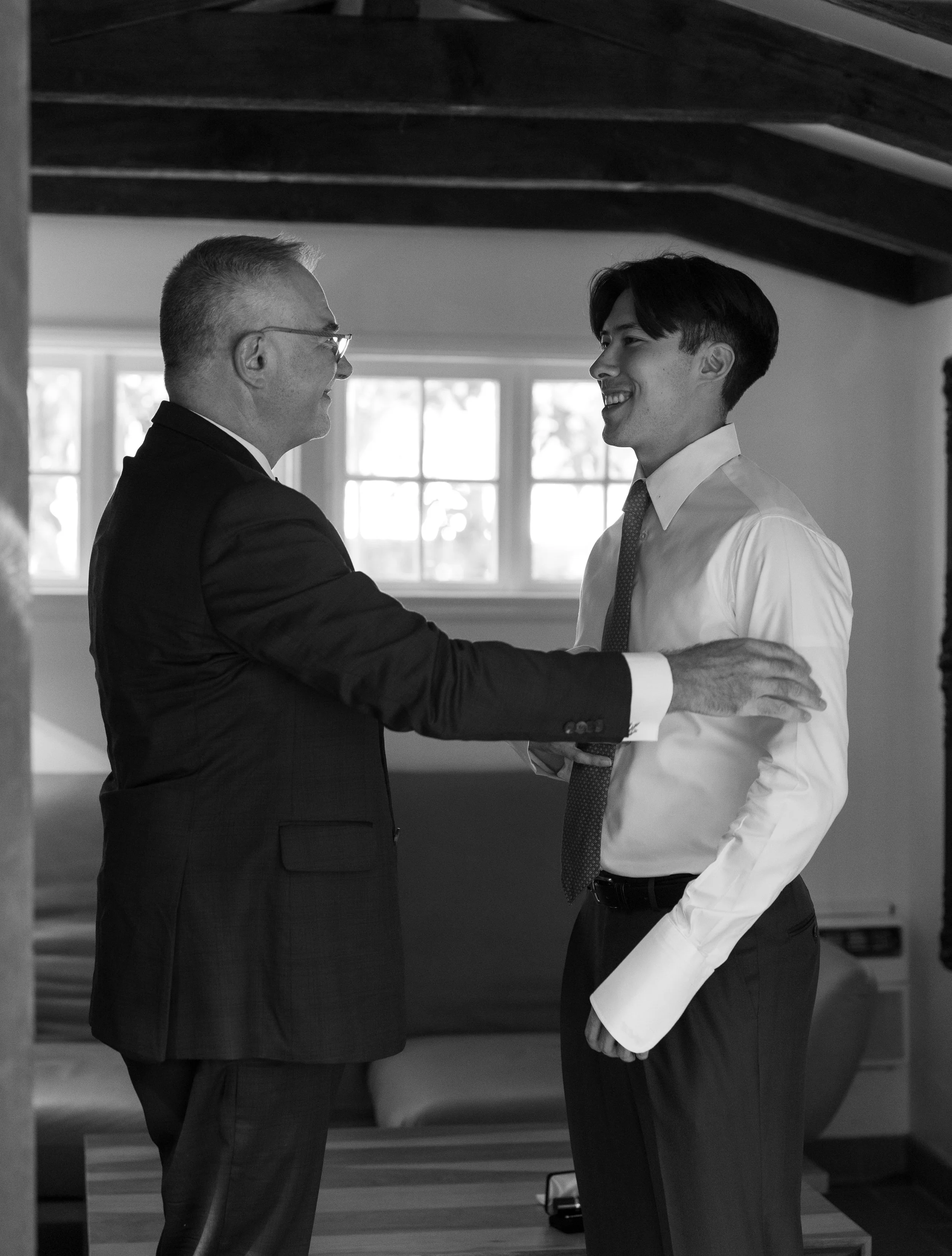 A groom and his father getting ready for a wedding, smiling and touching shoulders inside a room with windows, dressed in formal attire.
