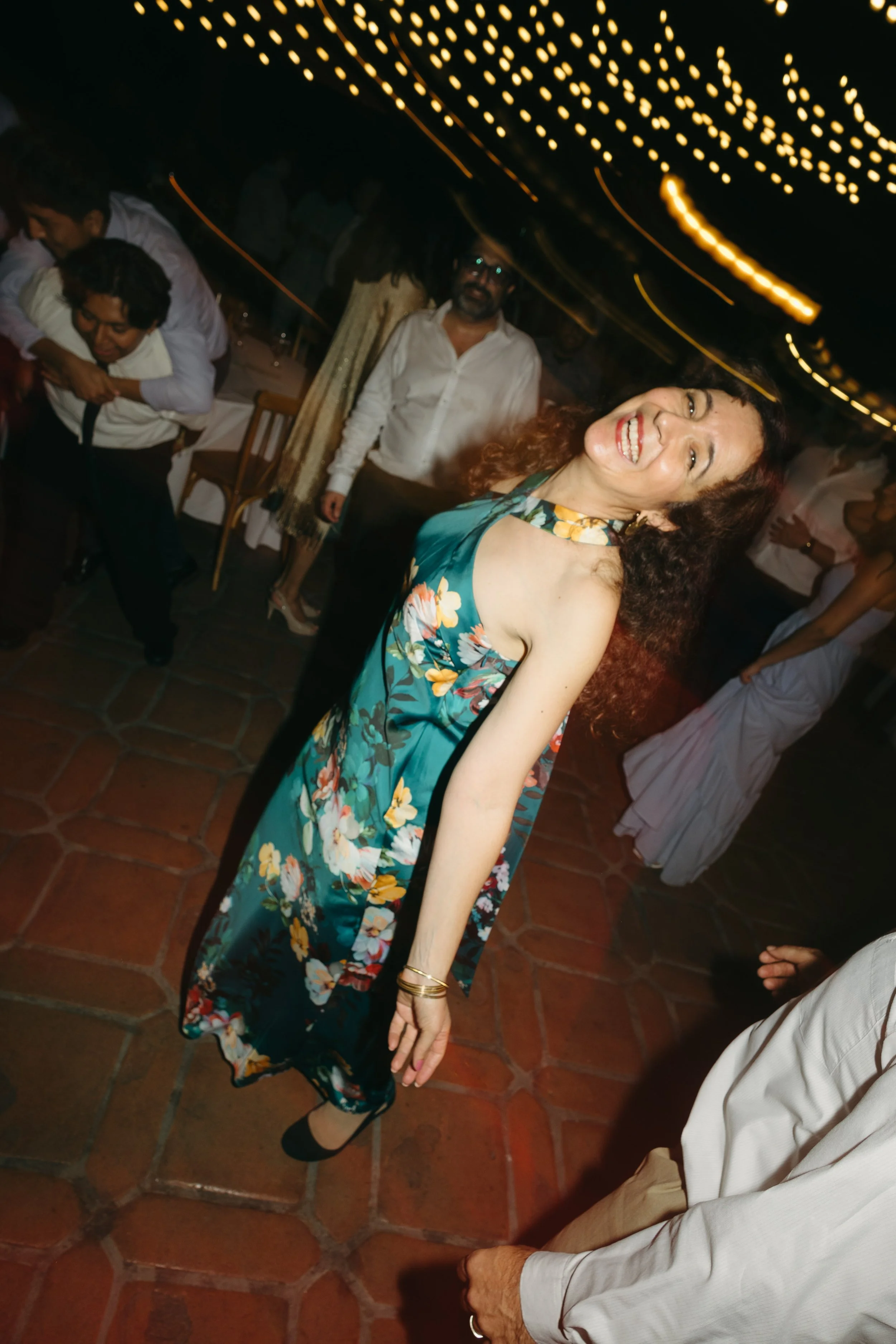 A woman in a floral halter dress dancing and smiling at a party with string lights overhead and people socializing in the background.