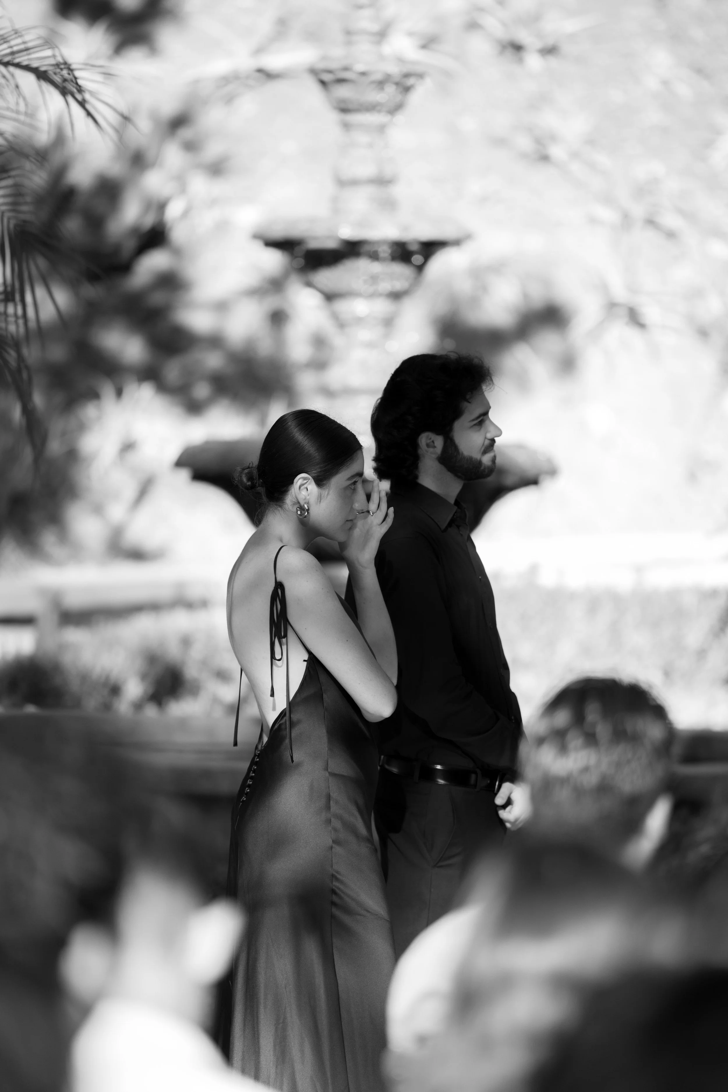A black and white photo of a woman and a man standing side by side outdoors in front of a fountain. The woman is whispering to the man, who is looking ahead calmly.