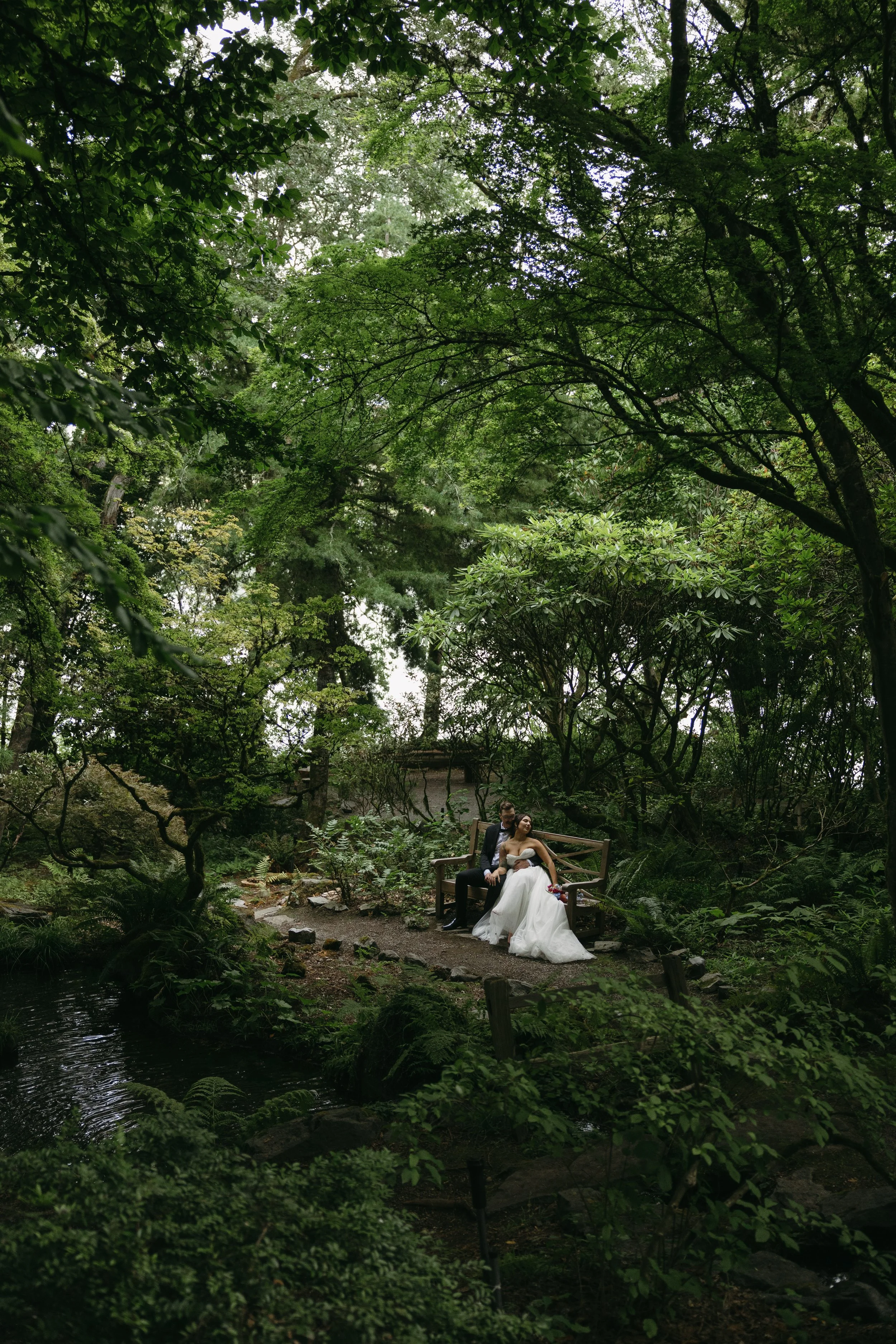 A bride and groom sitting on a wooden bench in a lush, green garden, surrounded by dense trees and foliage, with a small pond nearby at a Lakewold Gardens Wedding in Seattle