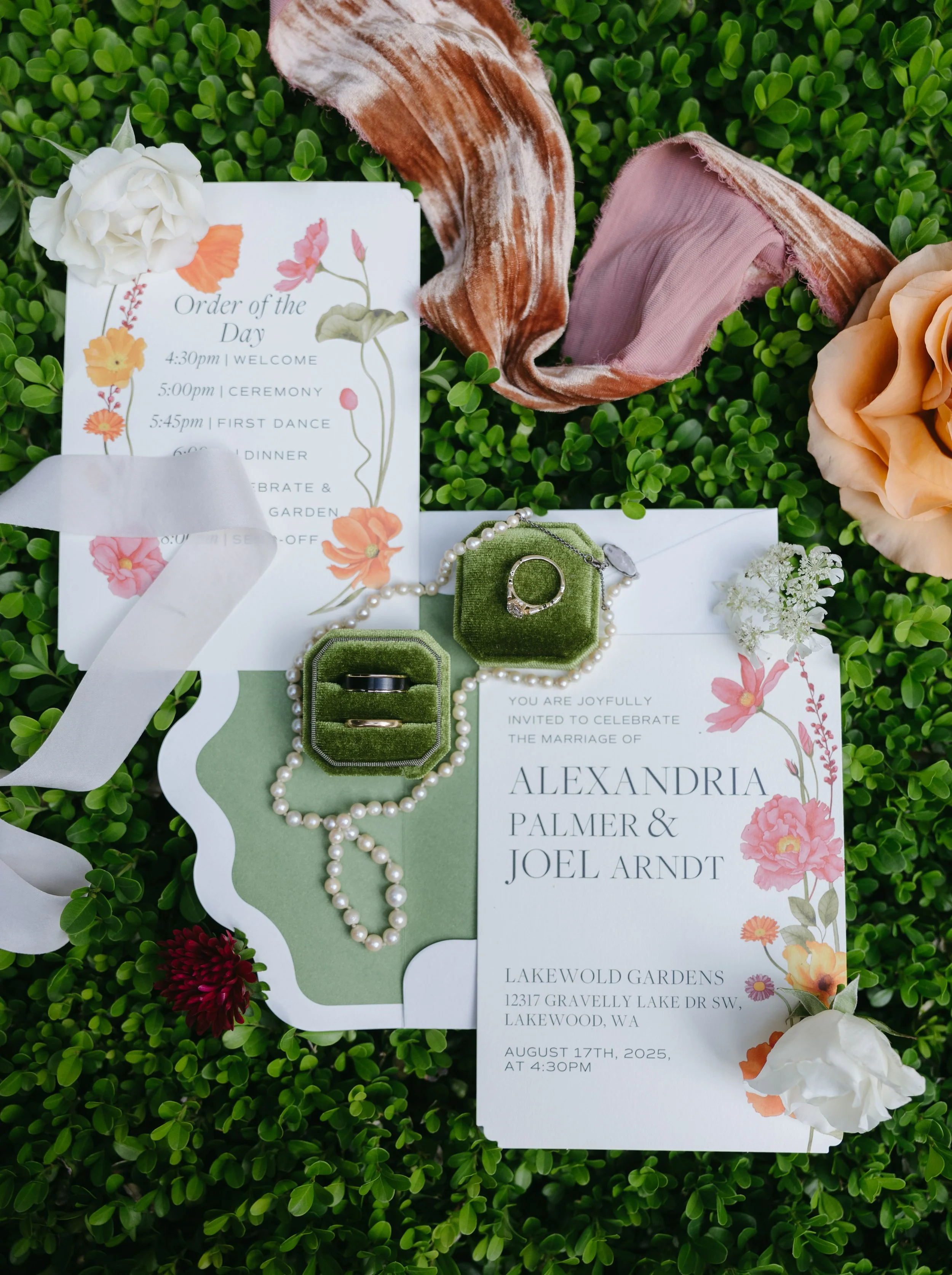 Wedding invitation and details, wedding rings in velvet boxes, pearl necklace, floral accents, velvet ribbon, and roses on grass