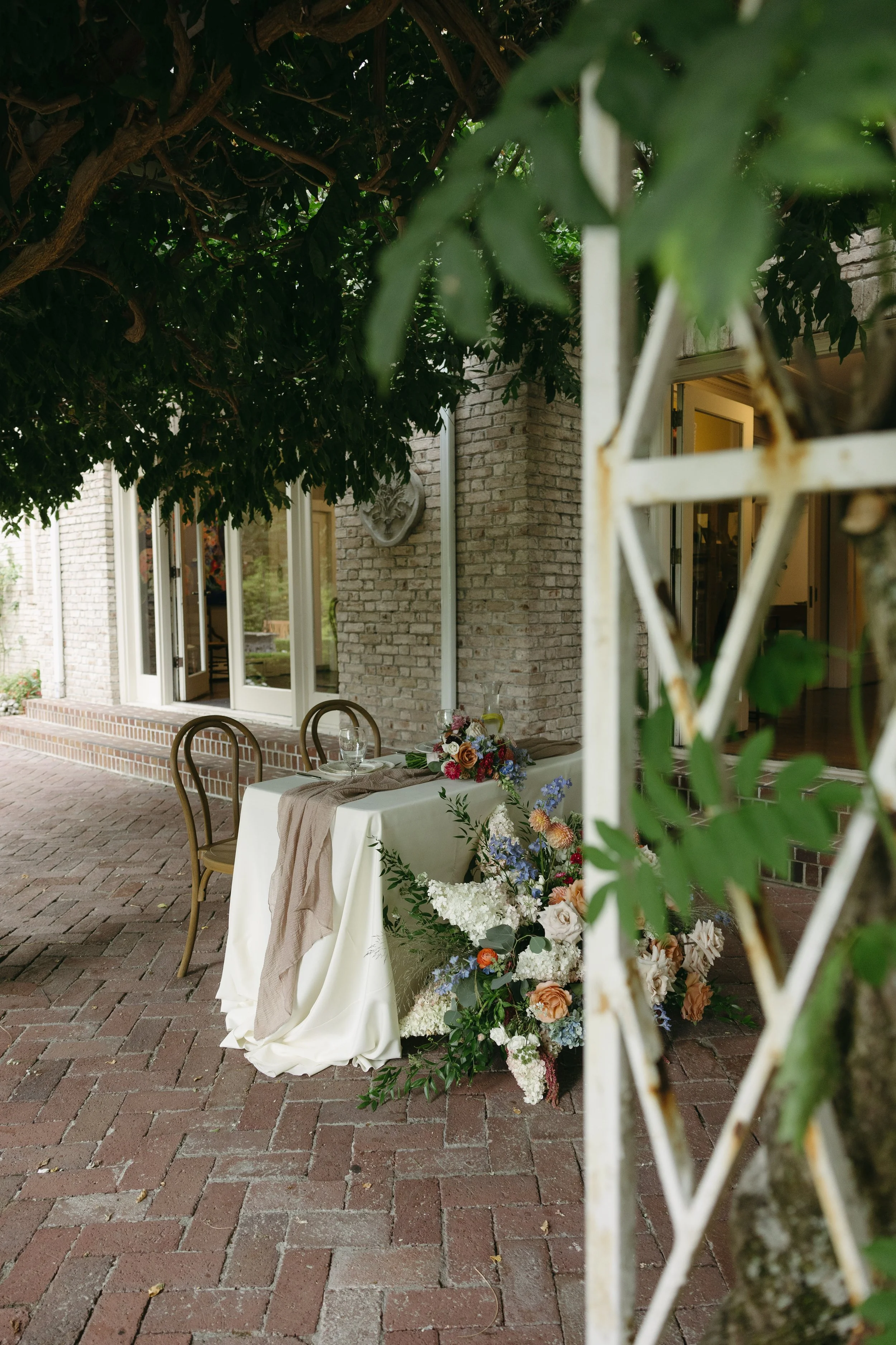 An outdoor table set for a meal with a white tablecloth, surrounded by elegant chairs, next to a building with brick walls and large windows, adorned with a colorful flower arrangement and greenery