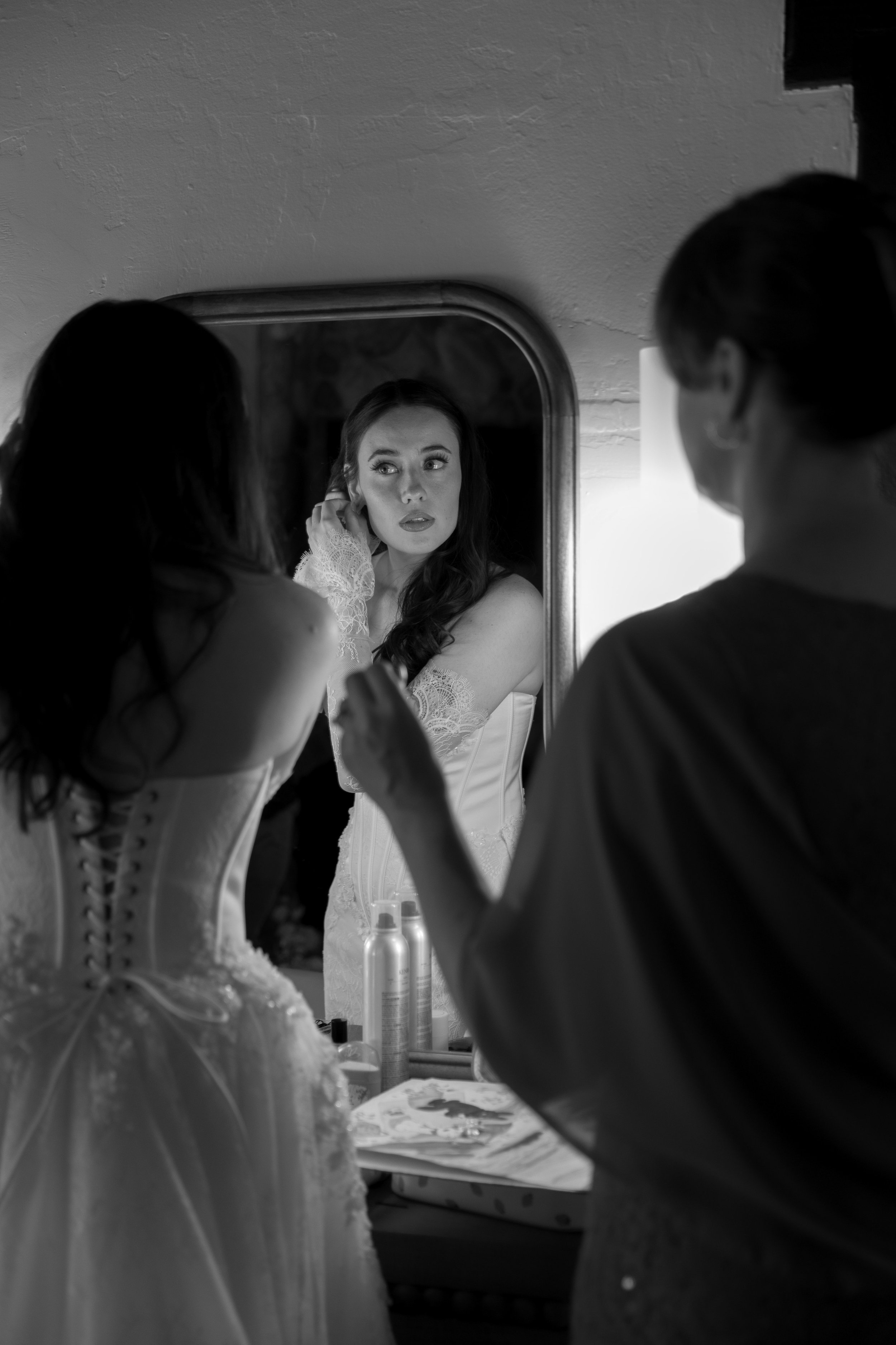 A bride and her mother are getting ready in front of a mirror. The bride is adjusting her earrings, and they are both dressed in wedding attire. The scene is in black and white at a Rancho Las Lomas Wedding in Los Angeles